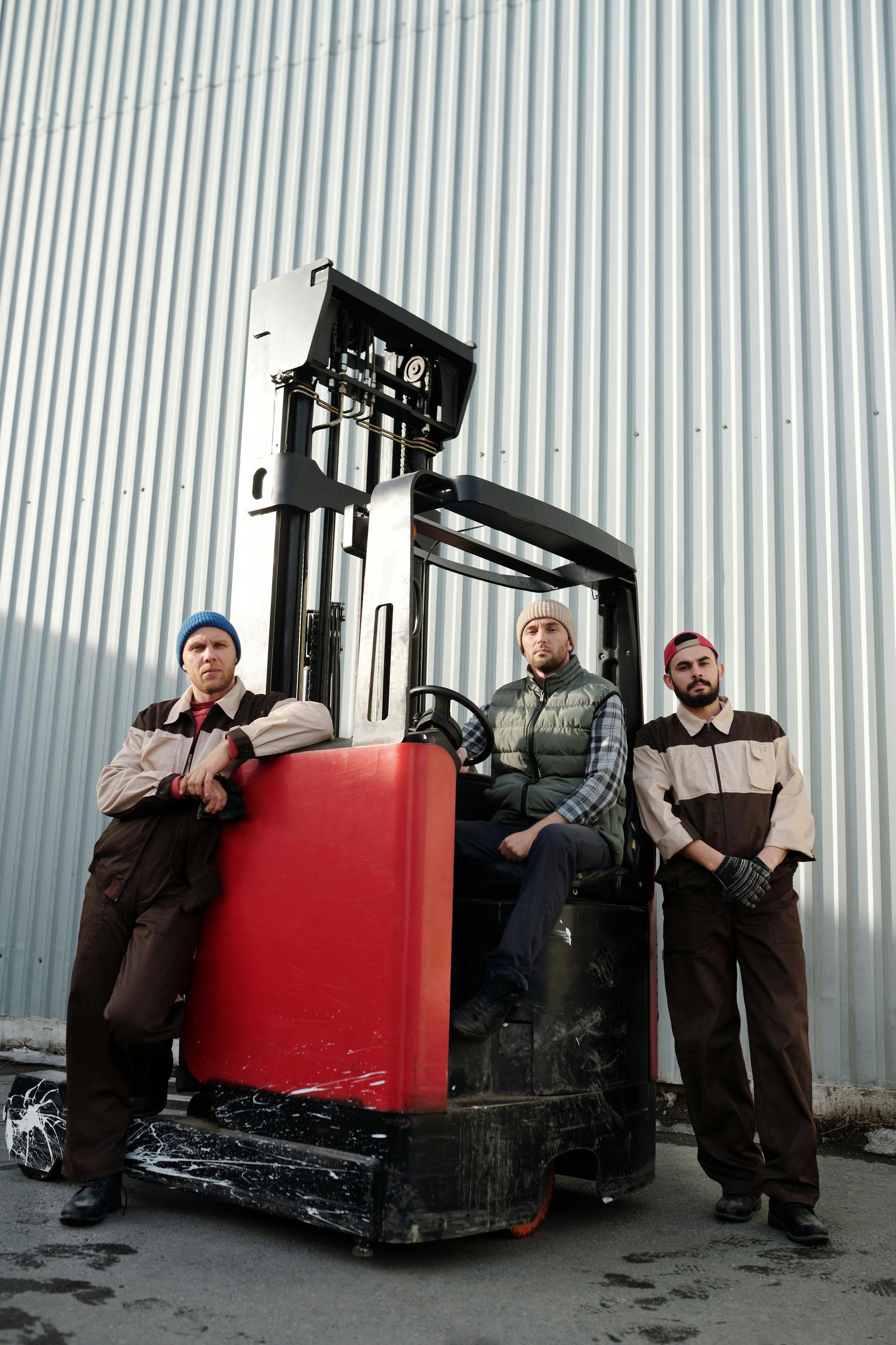 Three workers pose with a red forklift against a corrugated metal wall. They wear work clothes and hats.