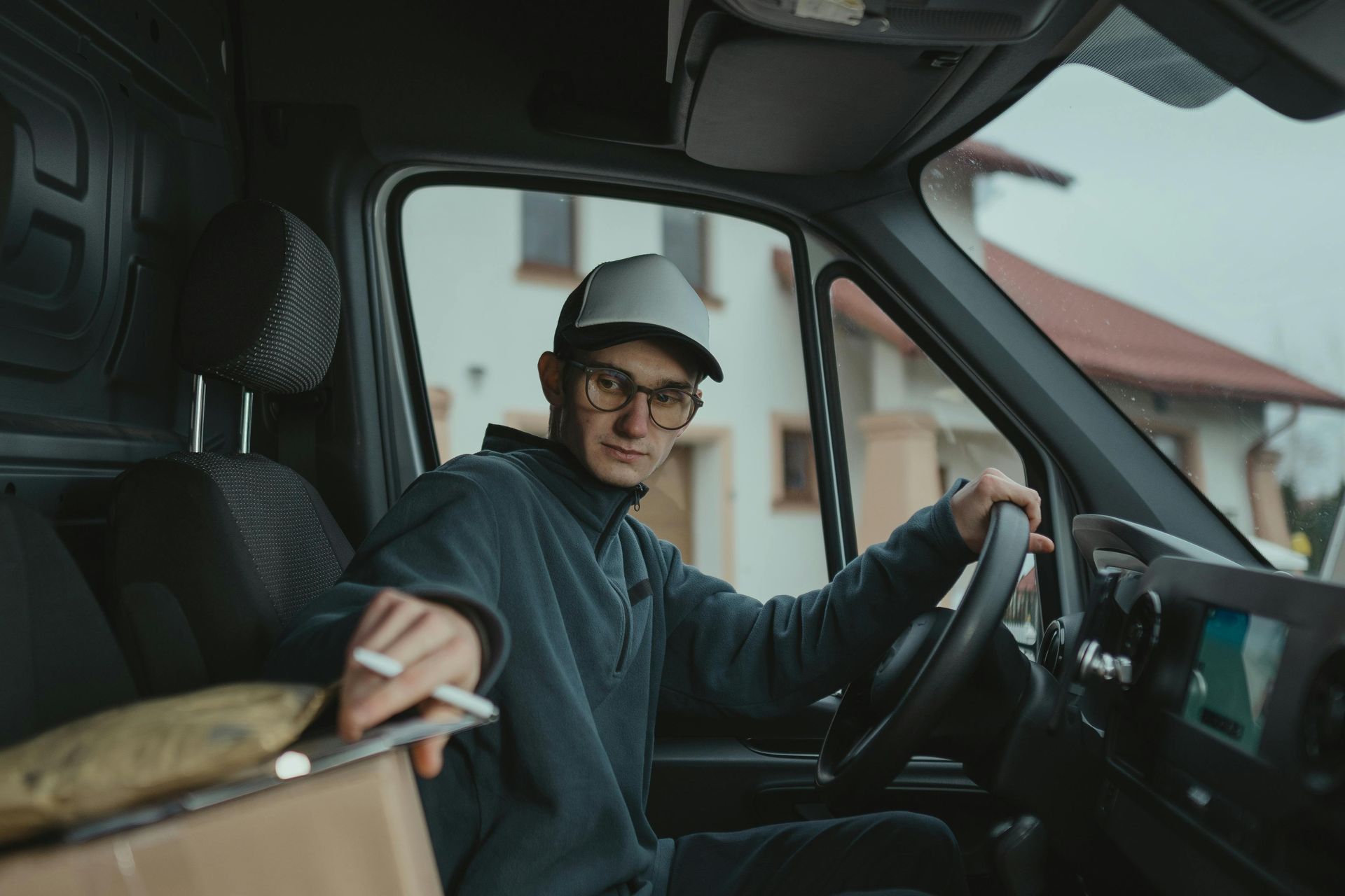 Man in delivery van, smoking cigarette, wearing cap and glasses, looking at camera.