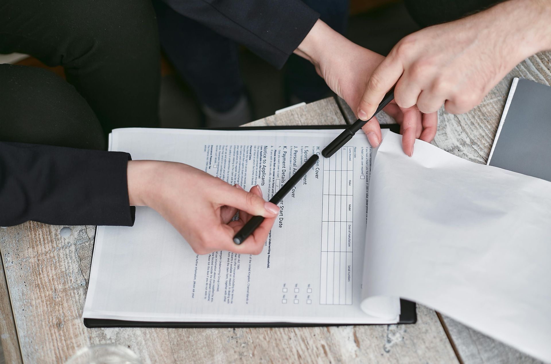 Hands pointing to a document on a table, reviewing details with pens, likely a business meeting.