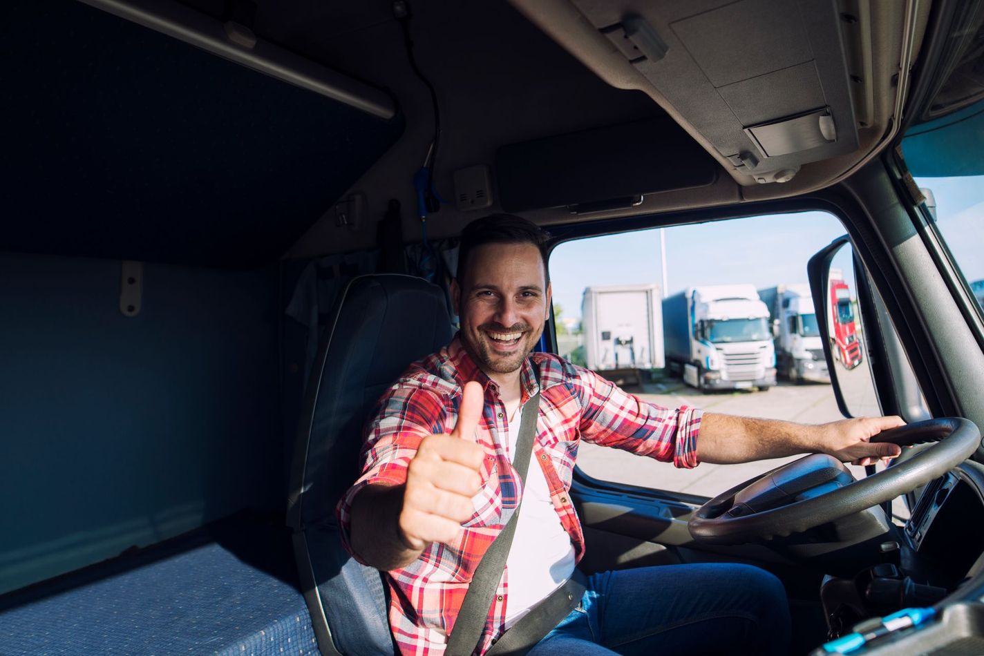 Smiling truck driver in a red plaid shirt giving a thumbs up, sitting in the driver's seat of a truck.