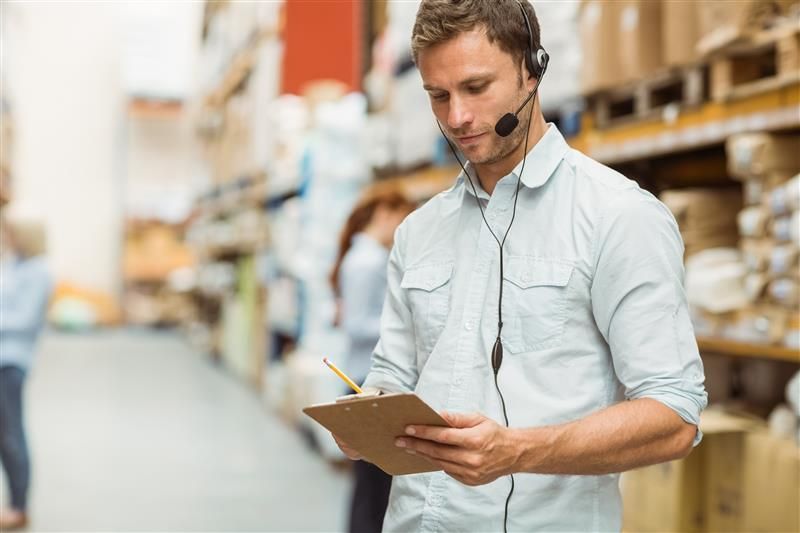 Warehouse worker with headset taking notes on a clipboard, surrounded by shelves of goods.