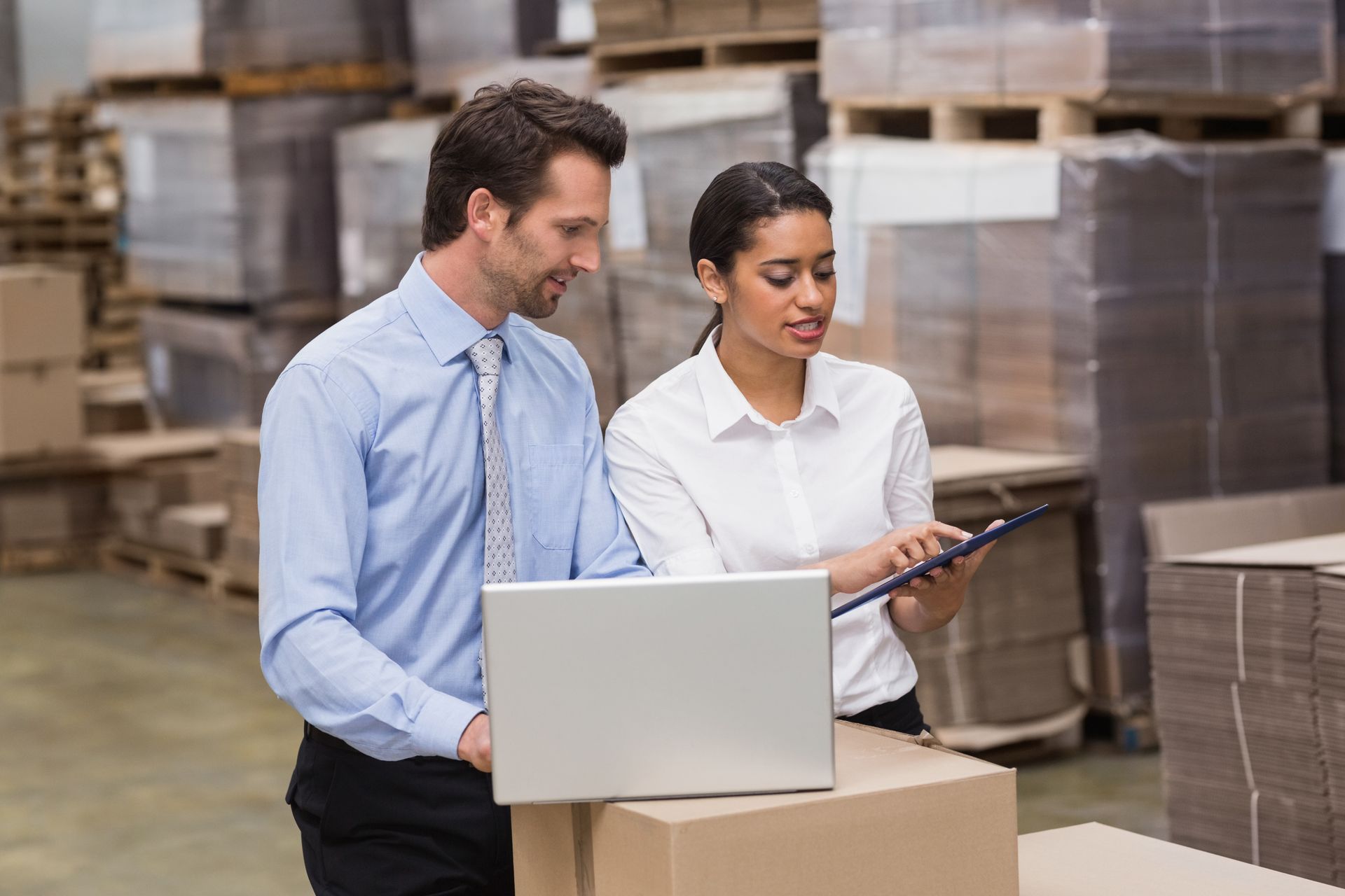 Man and woman looking at a laptop and clipboard in a warehouse; boxes and pallets are in the background.