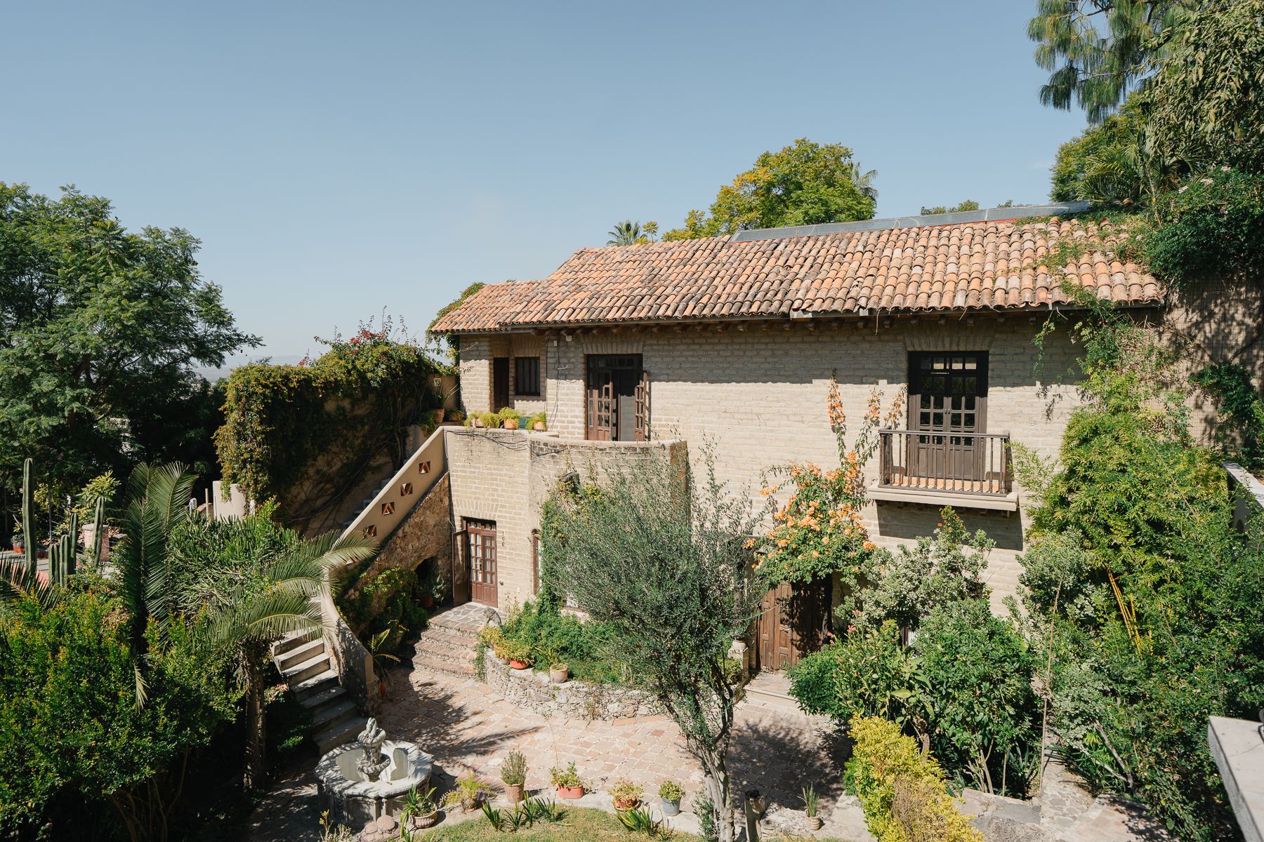 Edificio de piedra beige de dos pisos con techo de tejas de arcilla; rodeado de follaje verde y cielo azul.