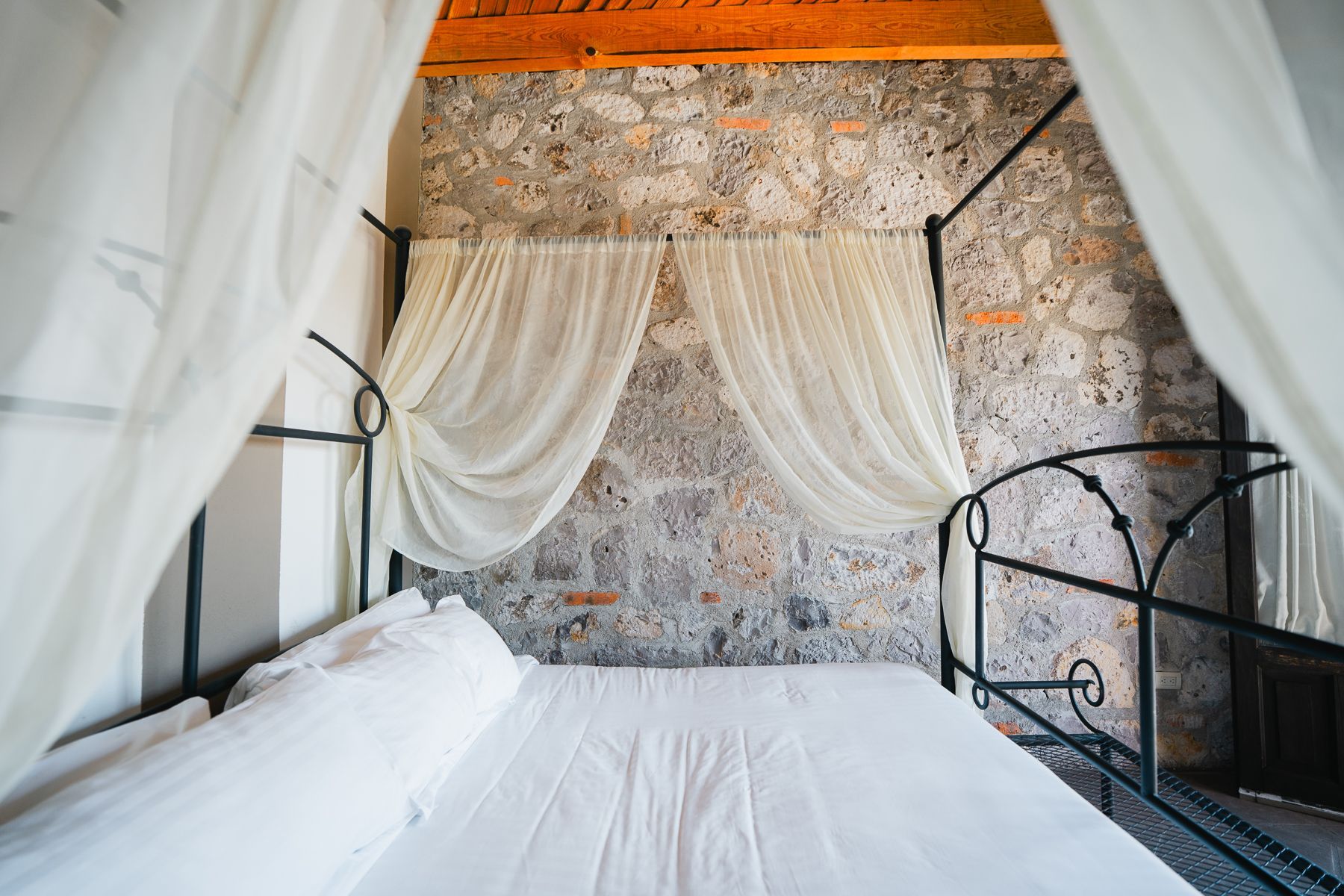 Cama con sábanas blancas y cortinas transparentes, frente a un muro de piedra.