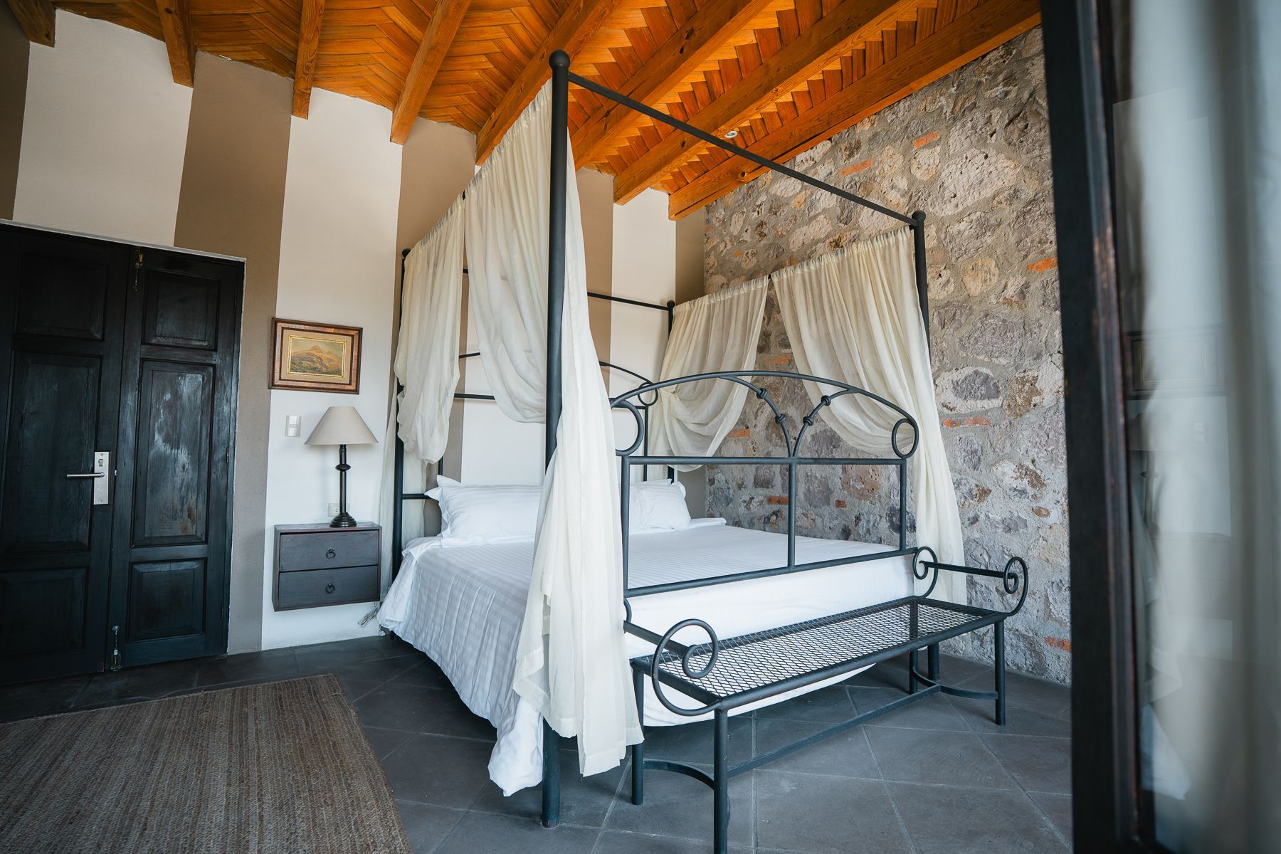 Dormitorio con cama con dosel, pared de piedra, techo de madera y puerta de madera oscura.