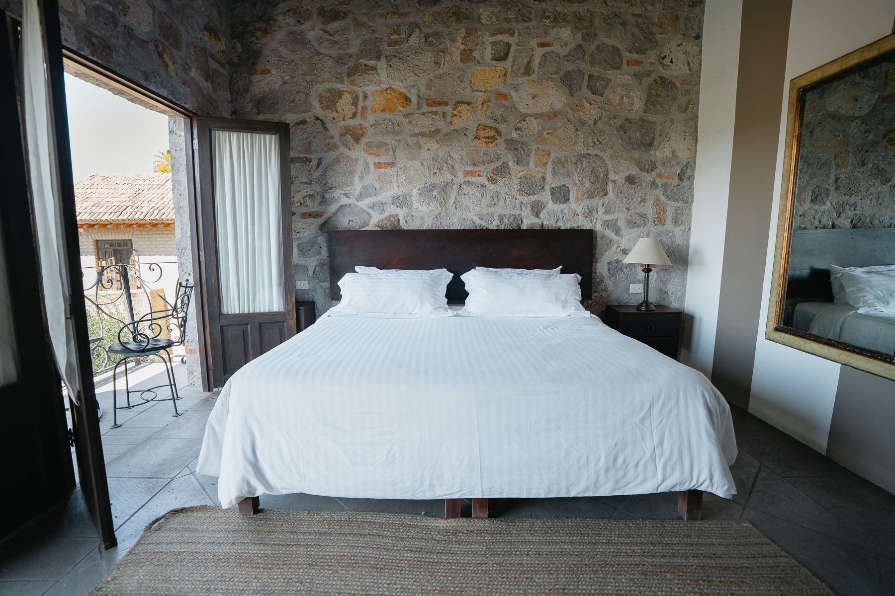 Dormitorio con pared de piedra, puerta abierta al balcón, cama con sábanas blancas y gran espejo.