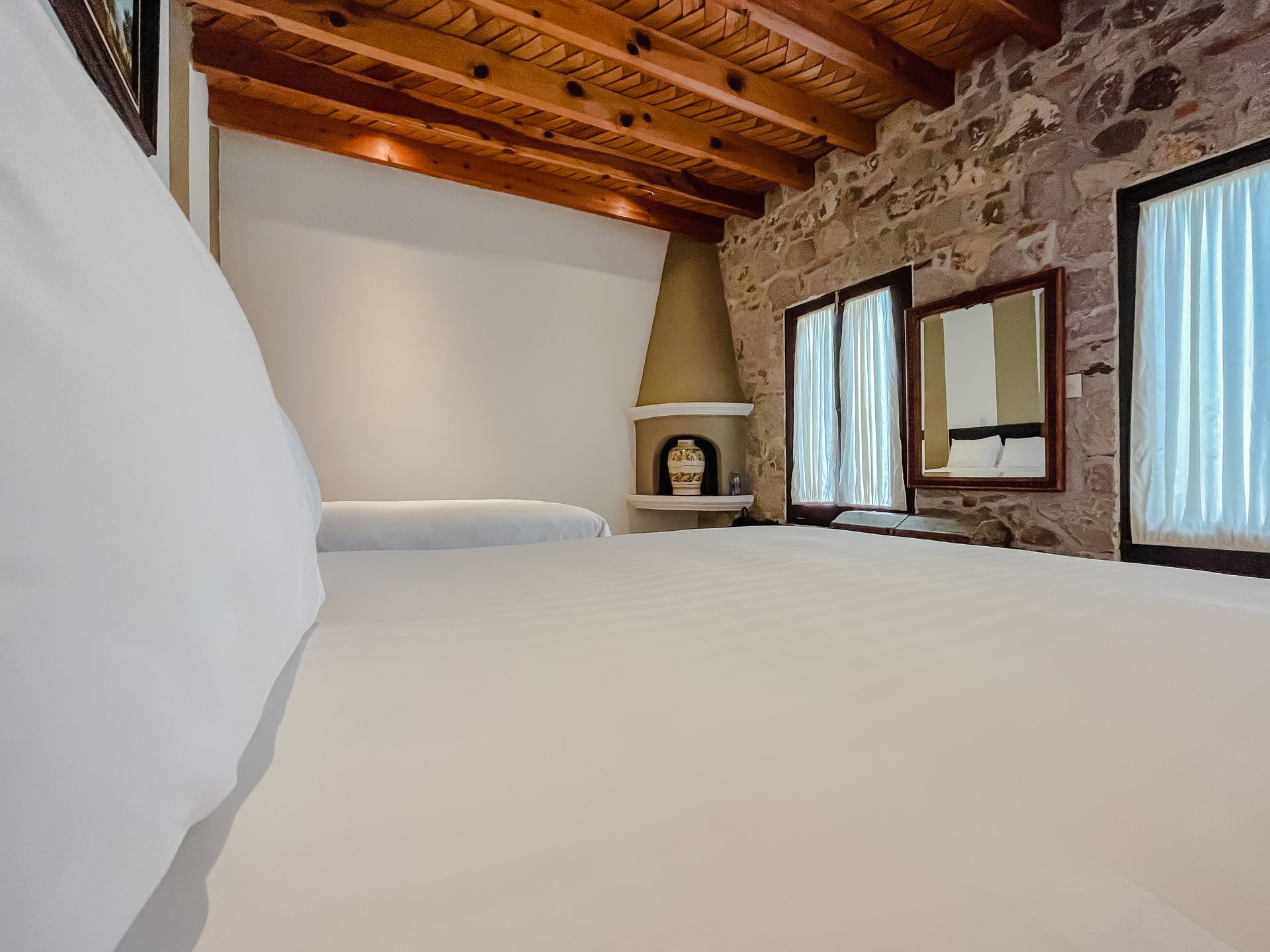 Dormitorio con cama blanca, paredes de piedra, techo de madera y ventanas con cortinas.