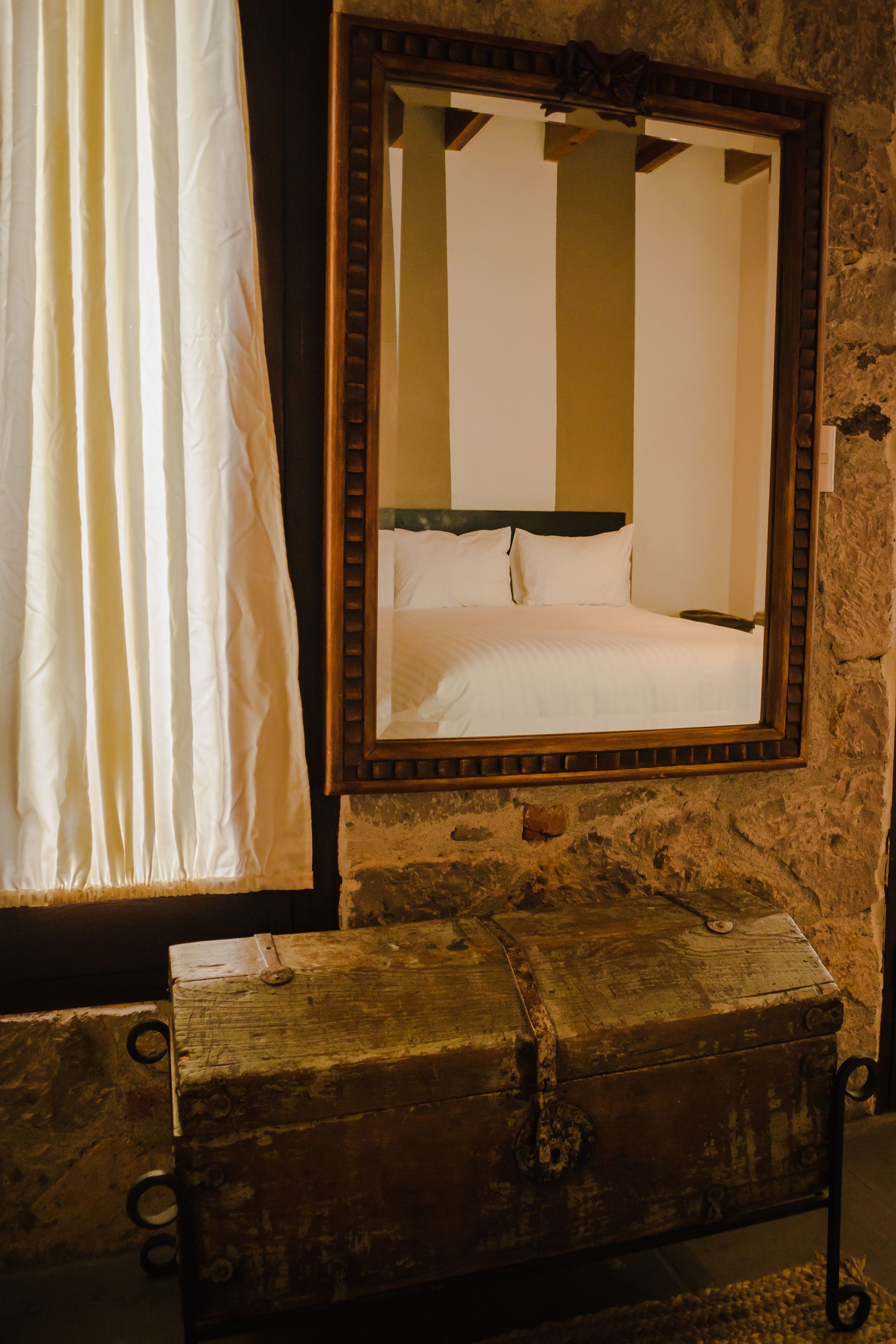 Baúl antiguo bajo un gran espejo que refleja una cama blanca. Cortina a la izquierda.