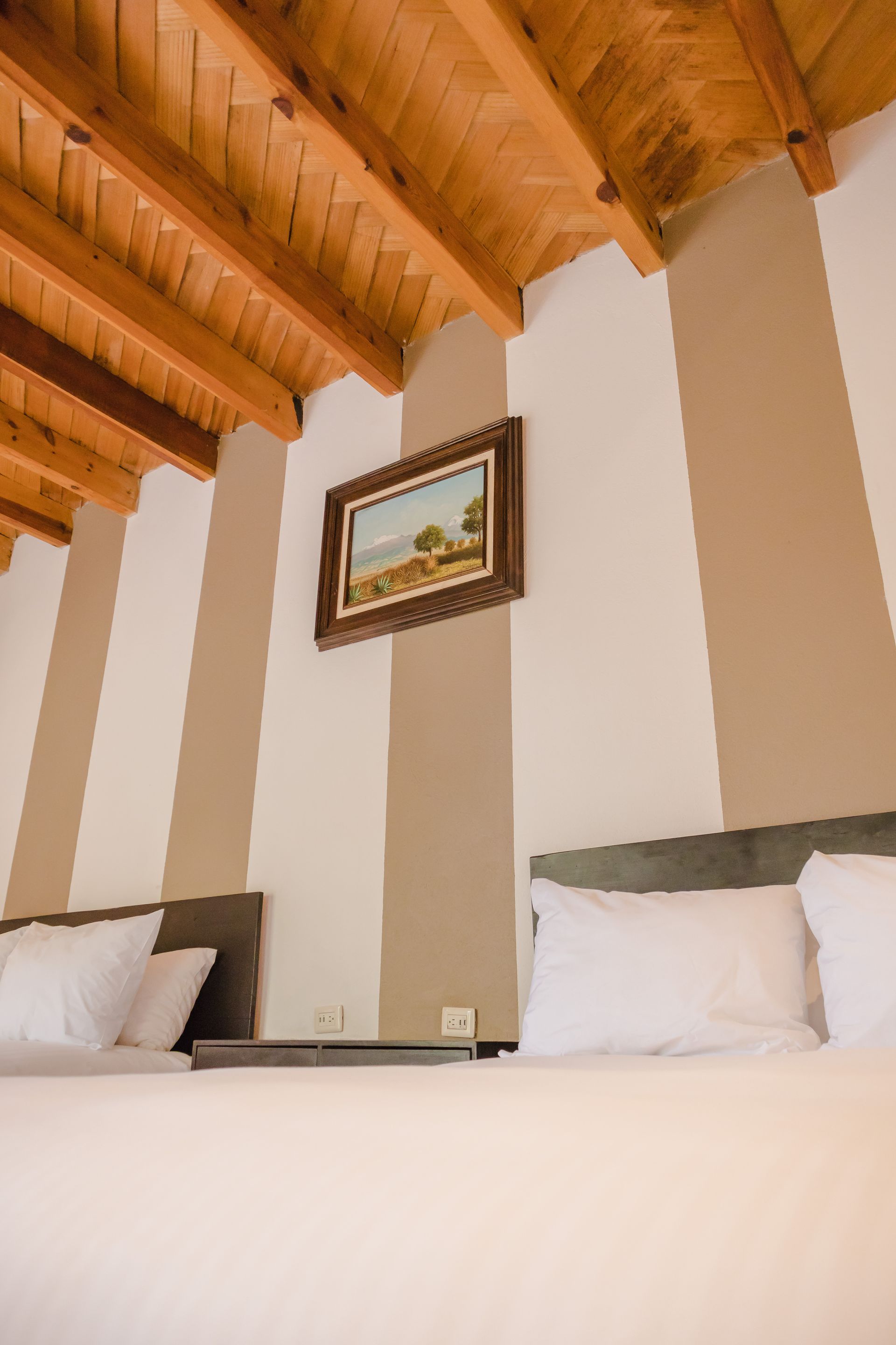 Dormitorio con paredes rayadas, techo de madera, dos camas y pintura de paisaje.