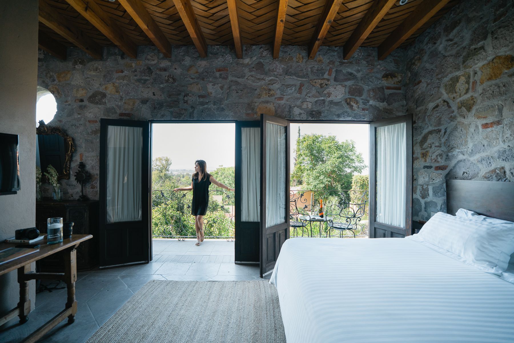 Dormitorio con paredes de piedra y puertas abiertas a un balcón con vistas a los árboles.
