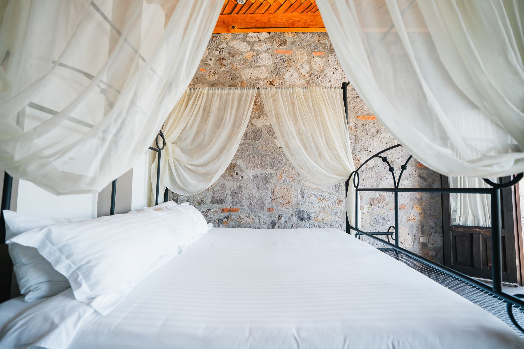 Cama con sábanas blancas y cortinas drapeadas contra una pared de piedra.