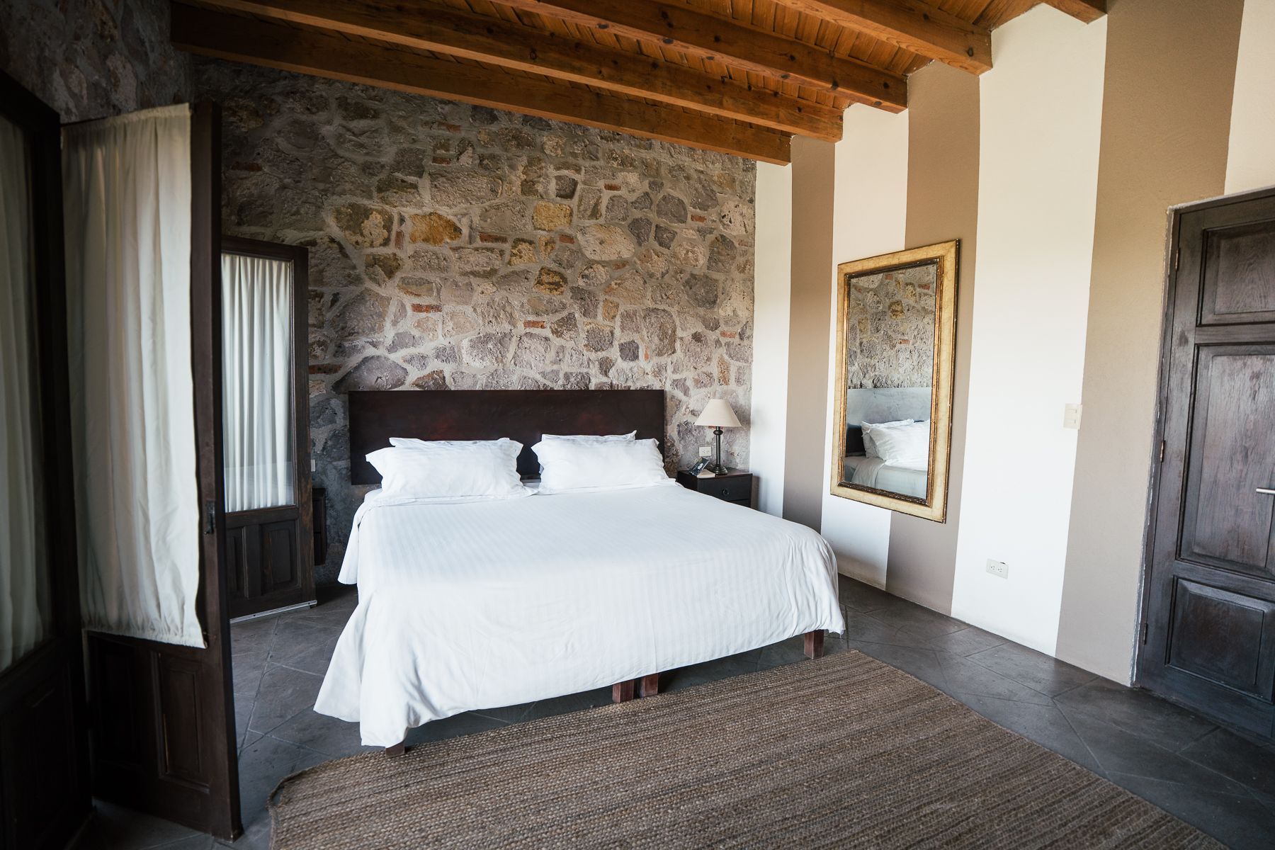 Dormitorio con pared de piedra y techo con vigas de madera. Cama con sábanas blancas, espejo grande y alfombra marrón.