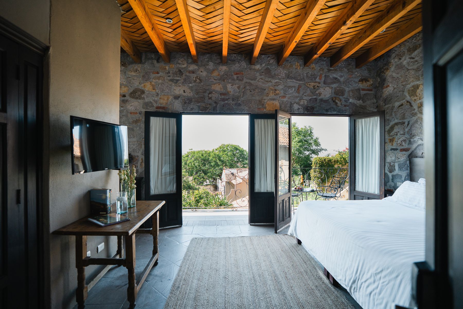 Dormitorio con paredes de piedra, techo de madera, cama, mesa y puertas abiertas a una vista.