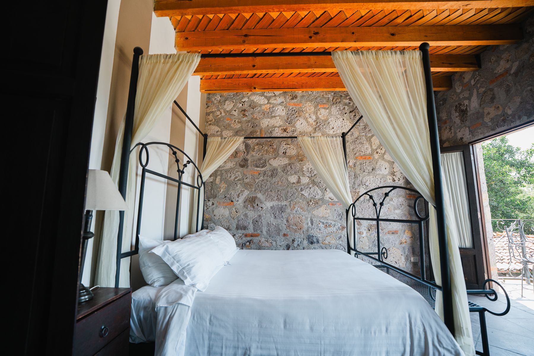 Dormitorio con cama con dosel; pared de piedra y techo de madera; cortinas transparentes.