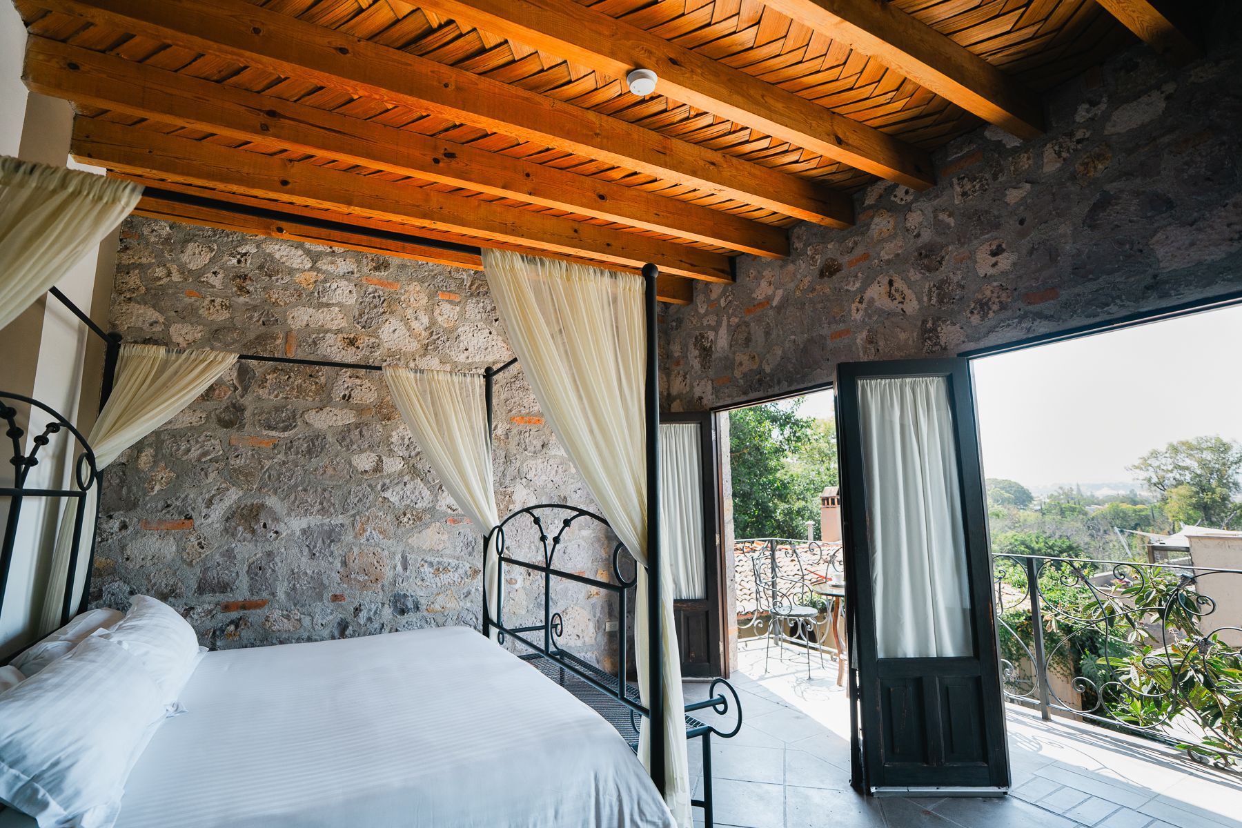 Dormitorio con paredes de piedra, cama con dosel con sábanas blancas y balcón con vista panorámica.