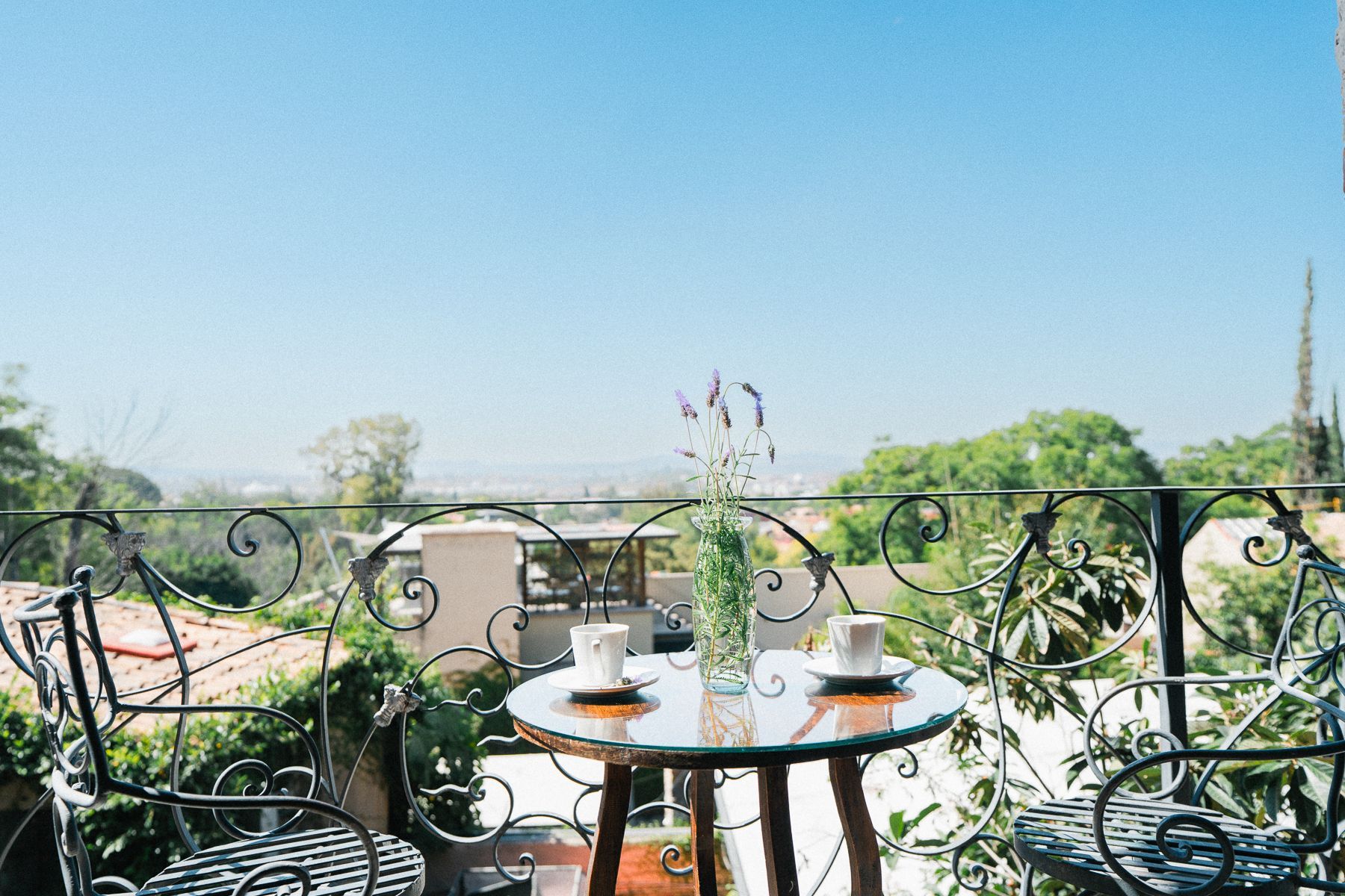 Balcón con mesa pequeña, dos tazas de café, arreglo floral, sillas de hierro forjado.