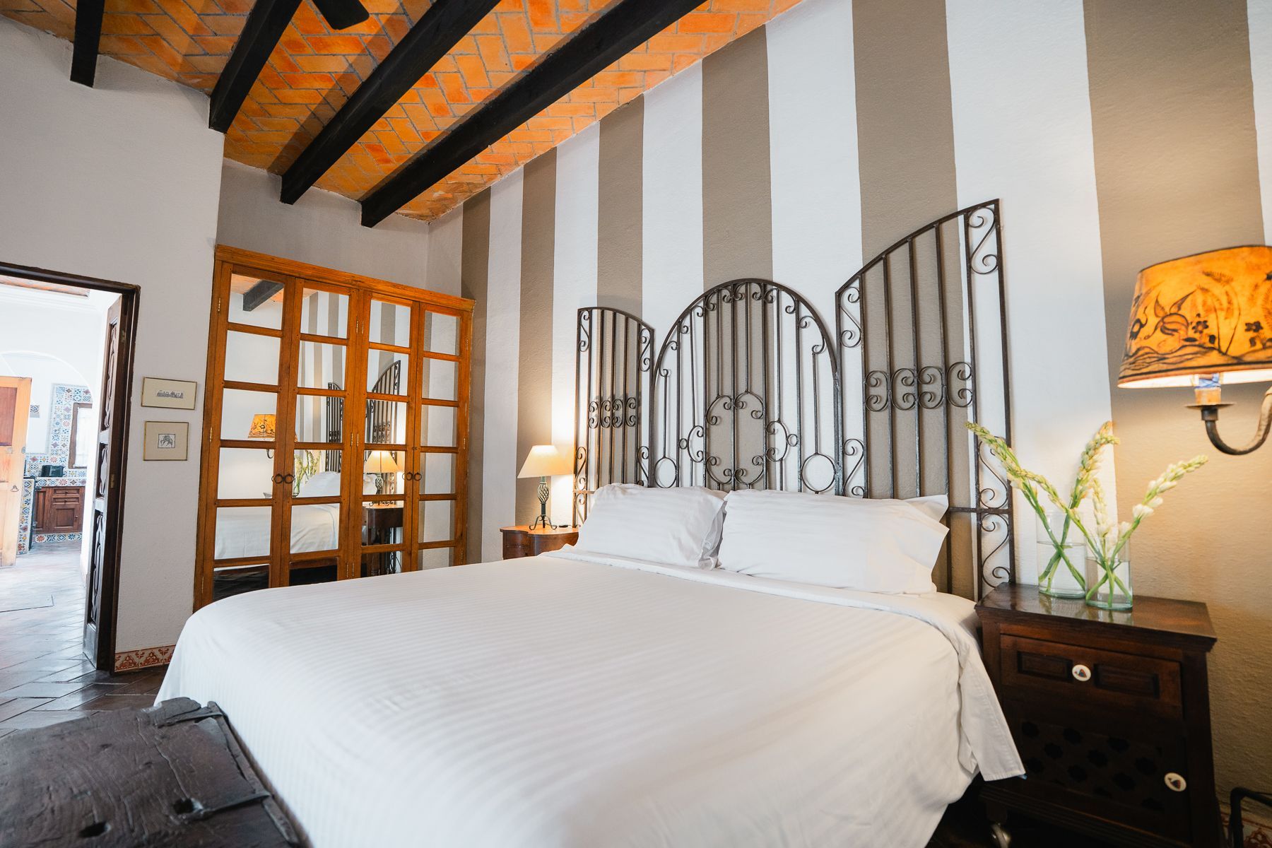 Dormitorio con cabecero de forja, pared rayada, vigas de madera en el techo y mesita de noche con lámpara.