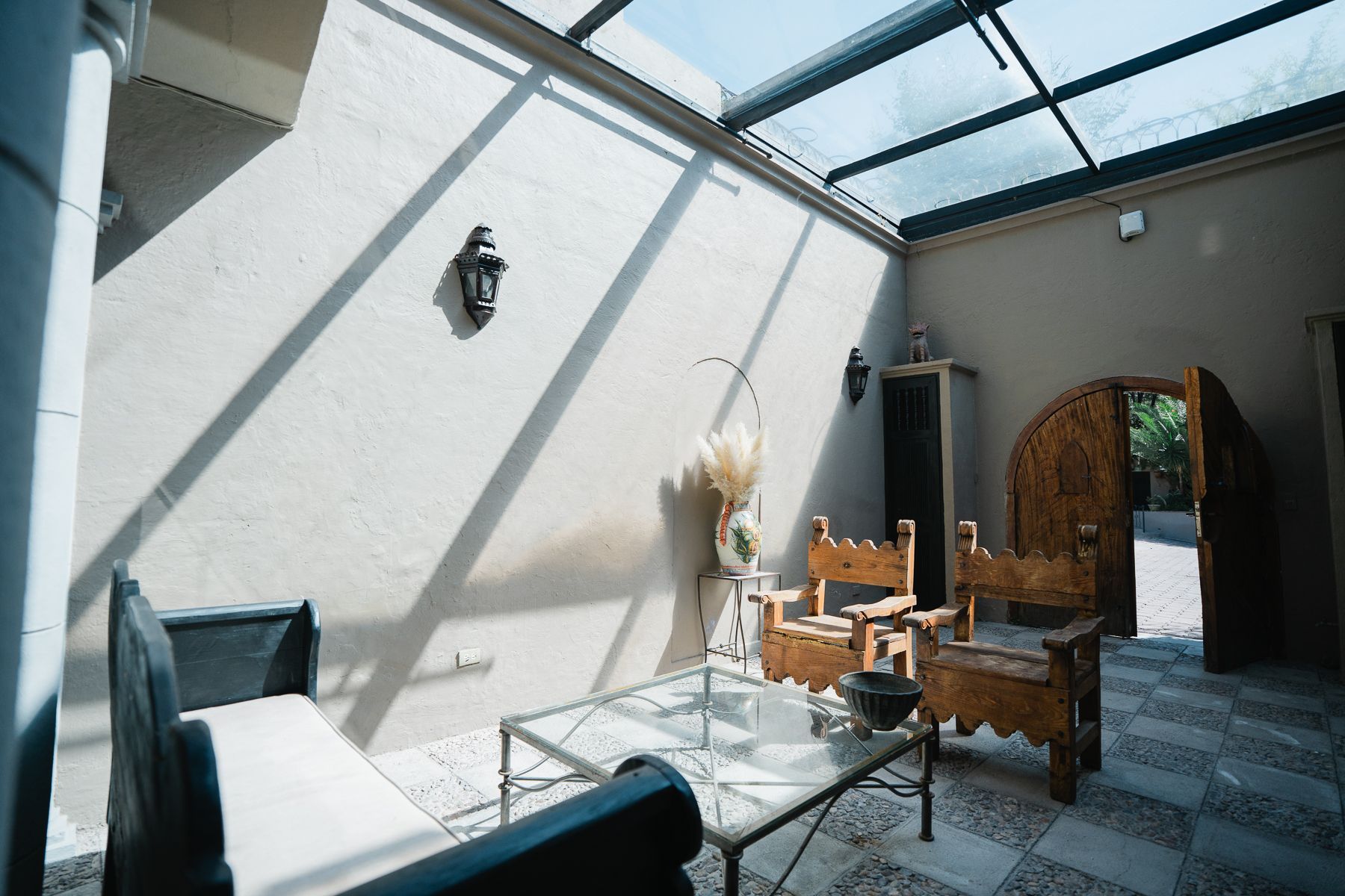 Patio con asientos, techo de cristal y puerta arqueada de madera. La luz del sol proyecta sombras en la pared blanca.