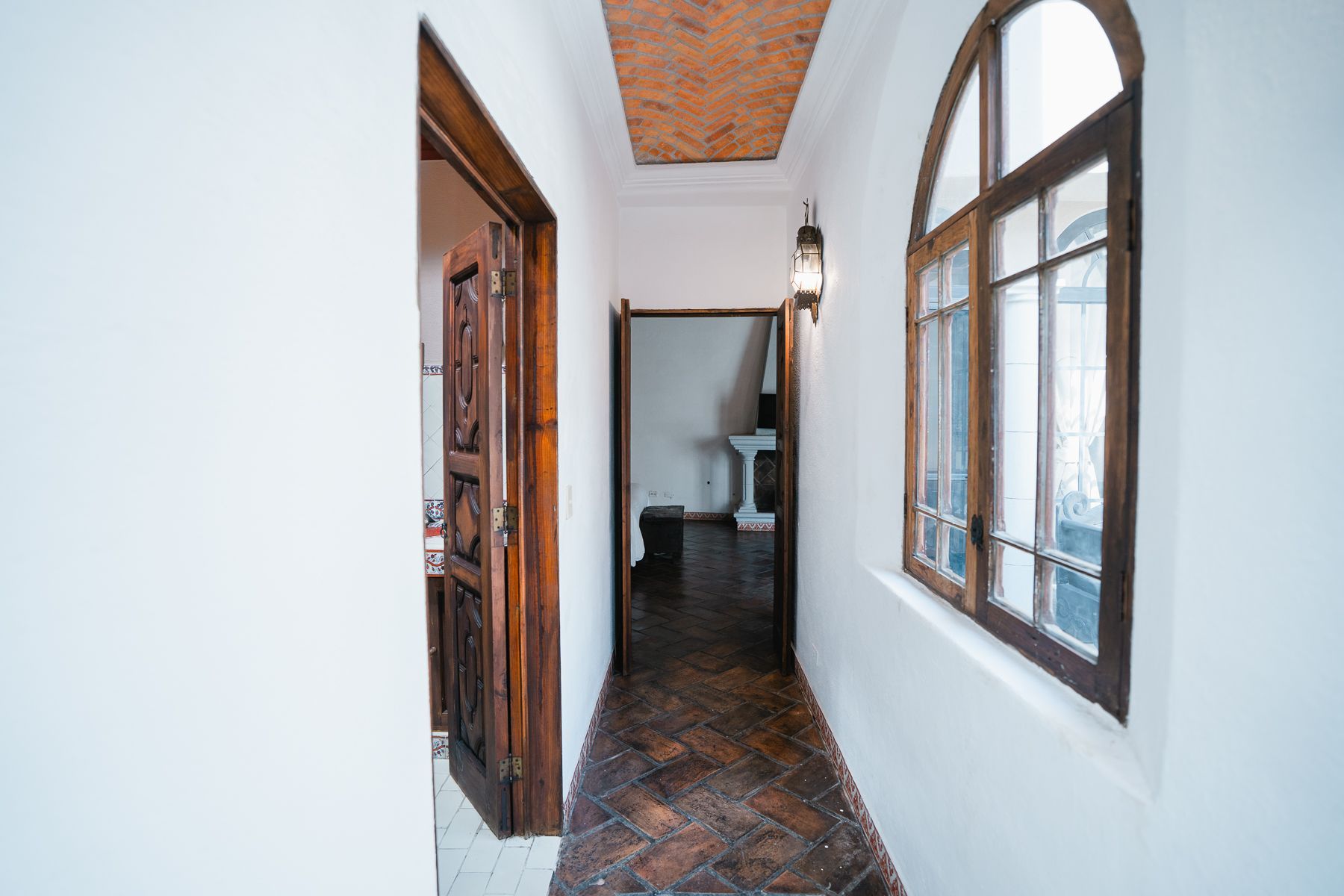 Pasillo estrecho con paredes blancas, ventana arqueada, puertas de madera oscura y suelo estampado.