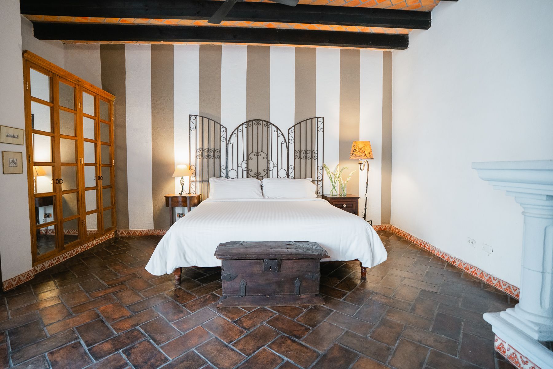 Dormitorio con pared rayada, cabecero de forja, suelo de madera y baúl a los pies de la cama.