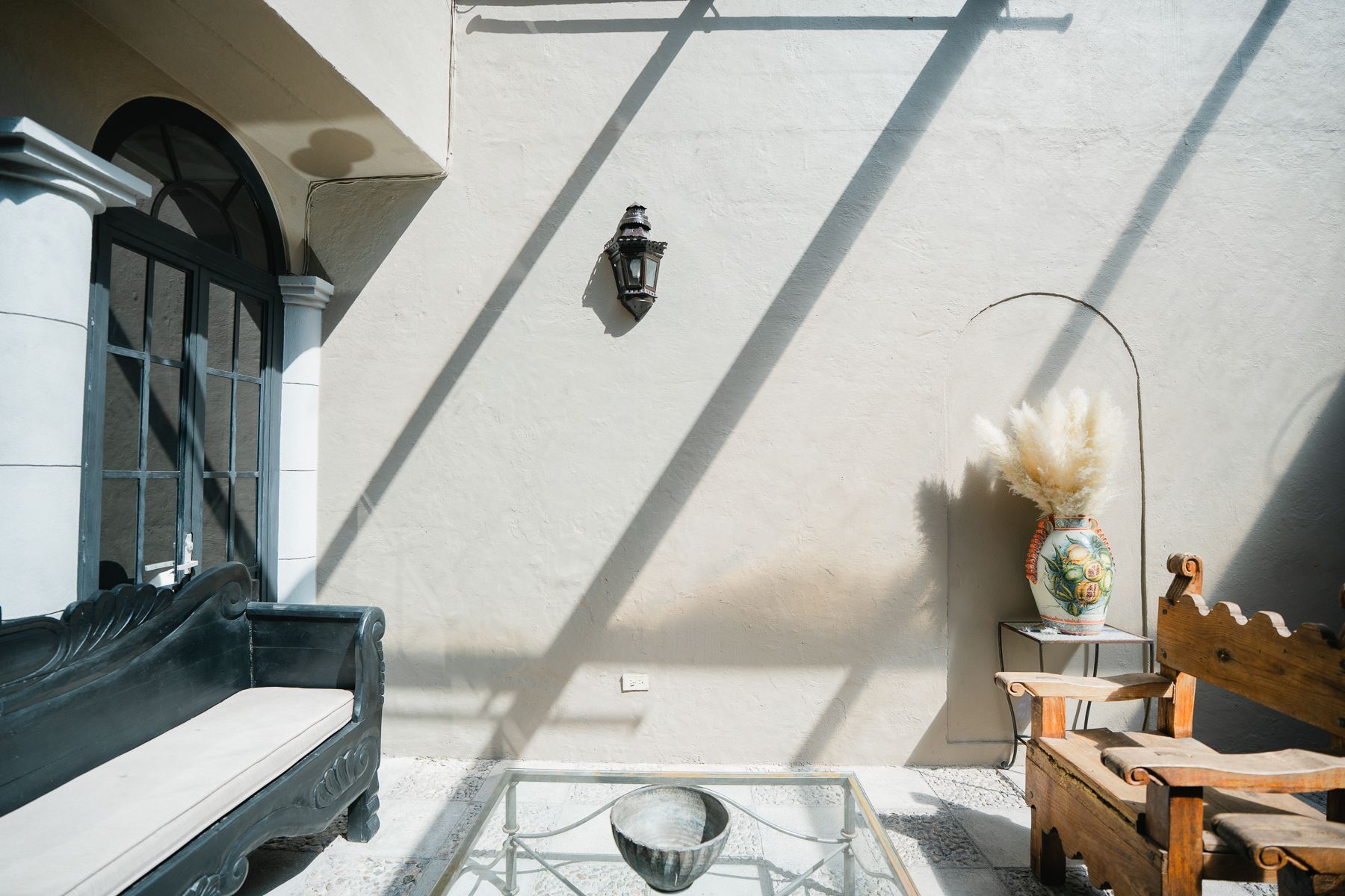 Patio exterior soleado con banco, silla y mesa de cristal. Diseños de sombras en la pared.