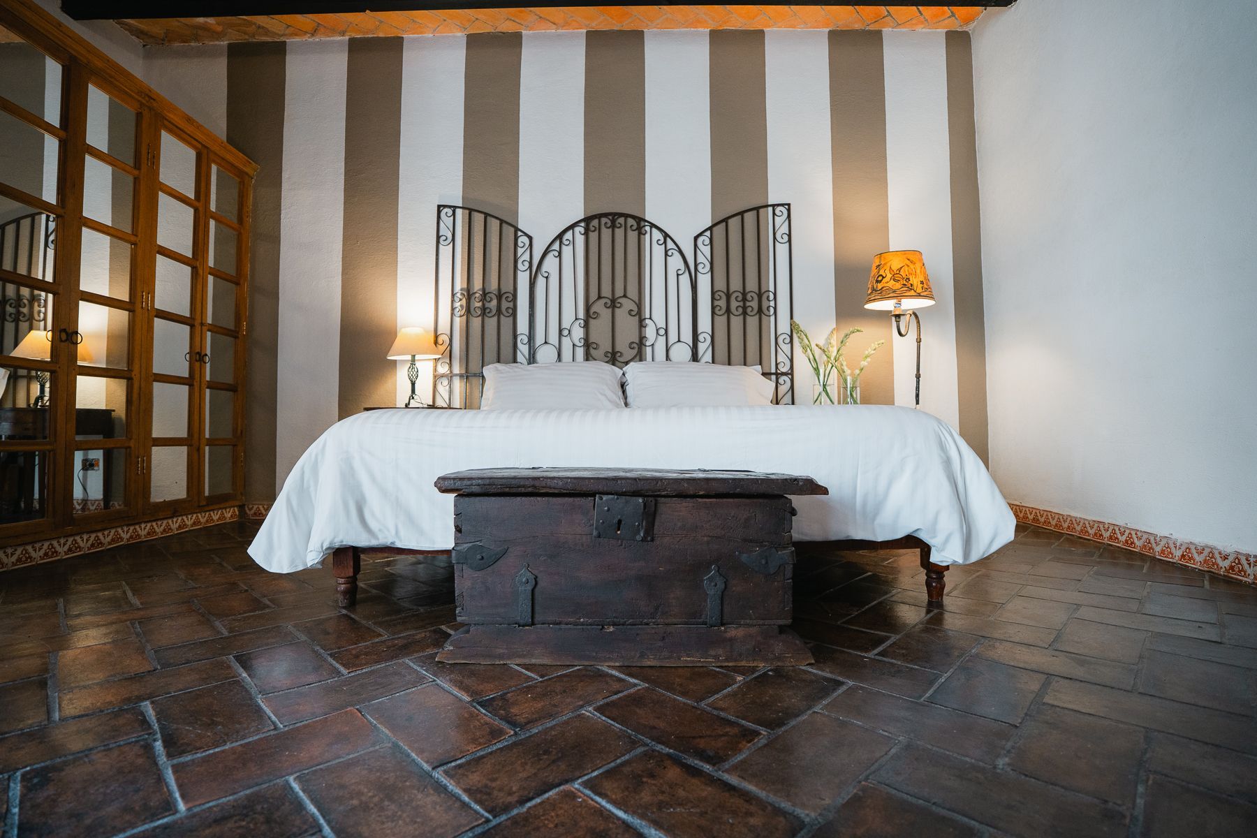 Dormitorio con cama, arcón oscuro, pared rayada, cabecero decorativo y suelo de madera.
