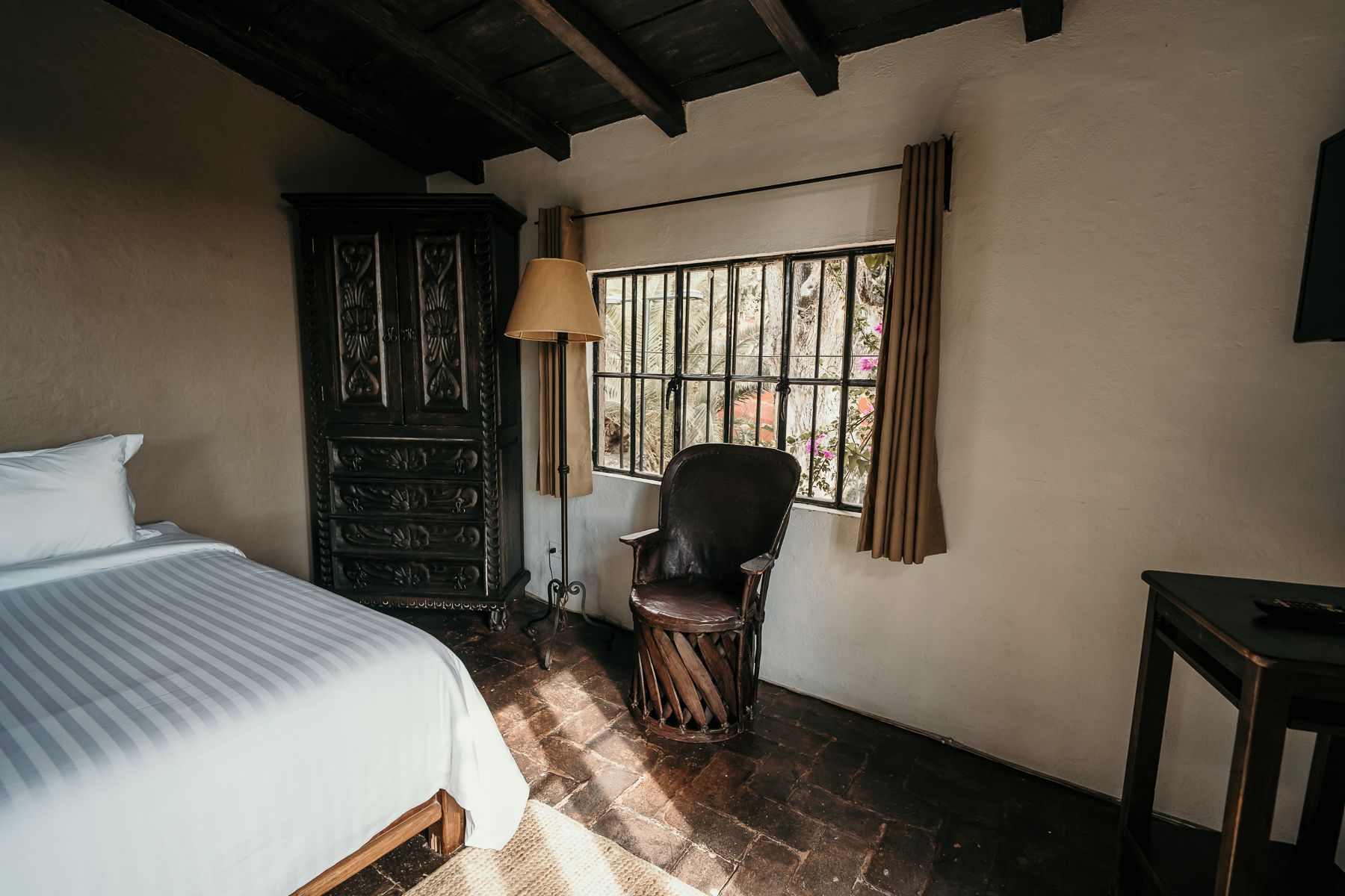 Dormitorio con cama, armario ornamentado, ventana, silla y lámpara de pie. La luz del sol entra a raudales por la ventana.