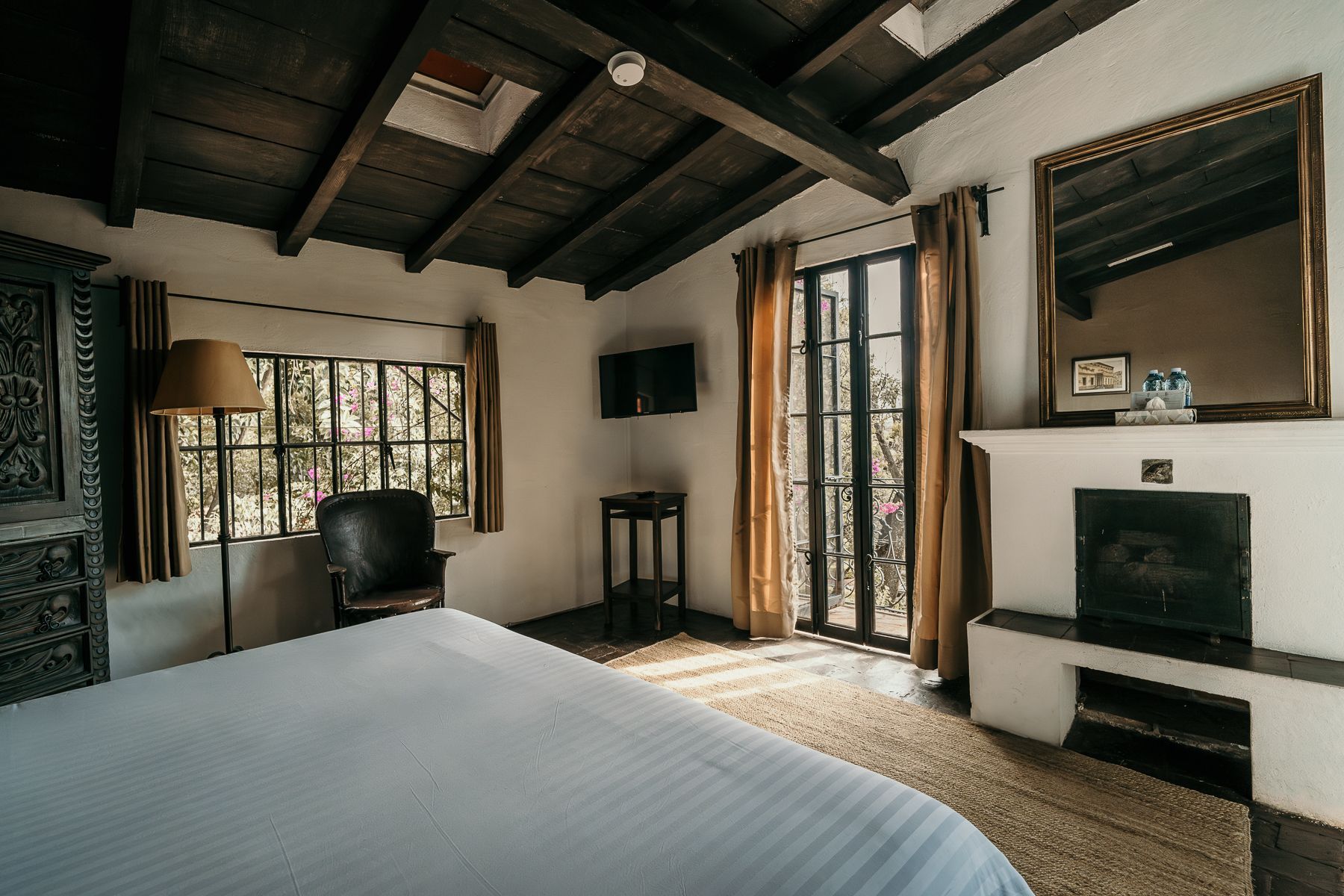 Dormitorio con paredes blancas, techo de madera oscura, ventanal, chimenea y cama.