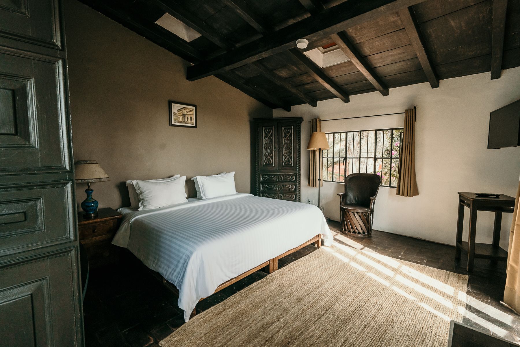 Dormitorio con cama blanca, muebles de madera oscura y luz natural que entra por una ventana.