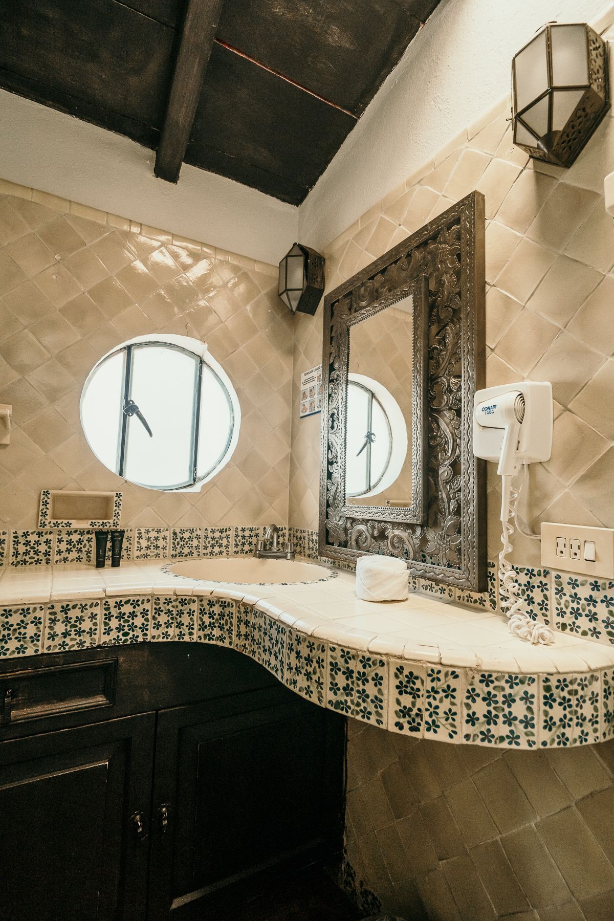 Baño con tocador de azulejos, espejo y ventana de ojo de buey. Muebles de madera oscura.