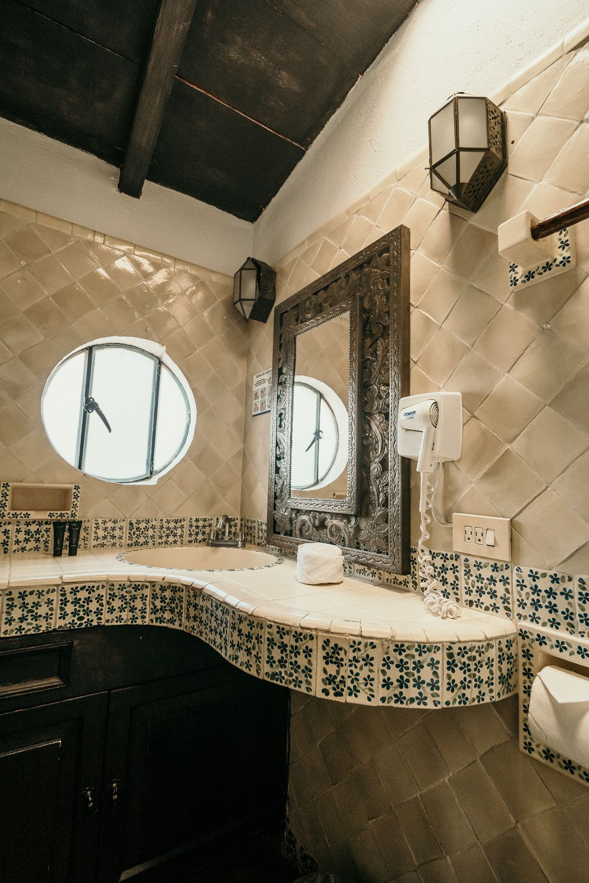 Baño con espejo ornamentado, azulejos decorativos, ventana ovalada, gabinetes de madera oscura y apliques de iluminación.