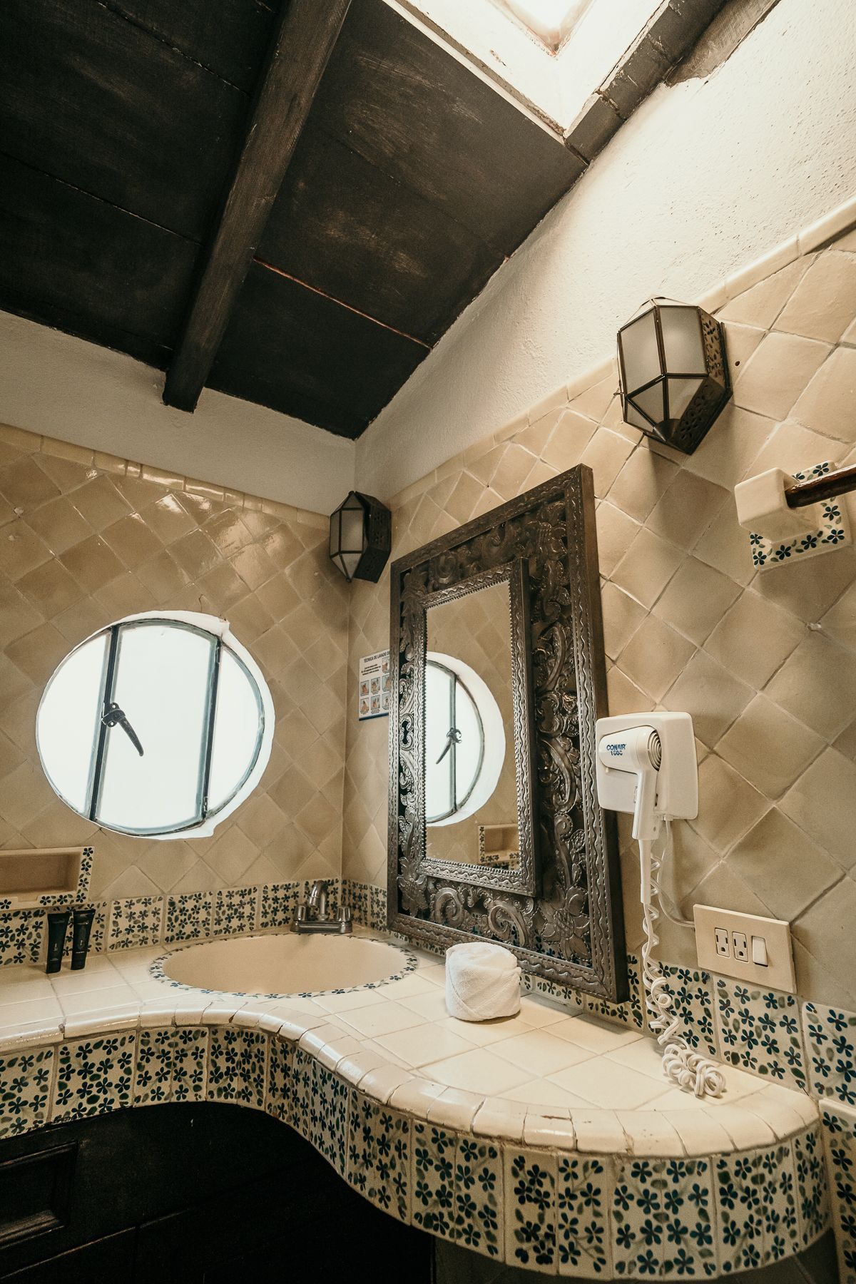 Baño con espejo ornamentado, azulejos decorativos, ventana circular y aplique de iluminación.