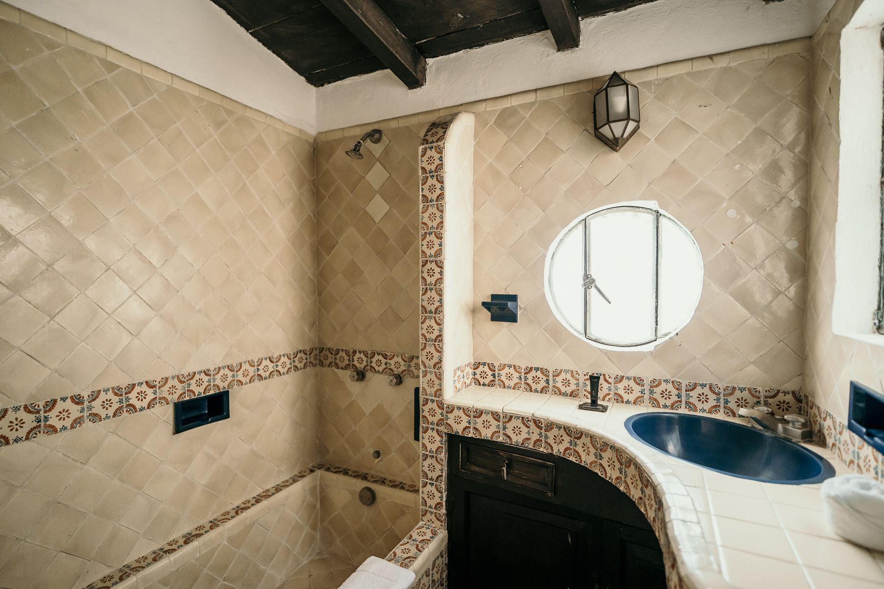 Baño con azulejos de color tostado con diseño de diamantes, lavabo azul, ventana redonda, vigas y molduras de madera oscura.