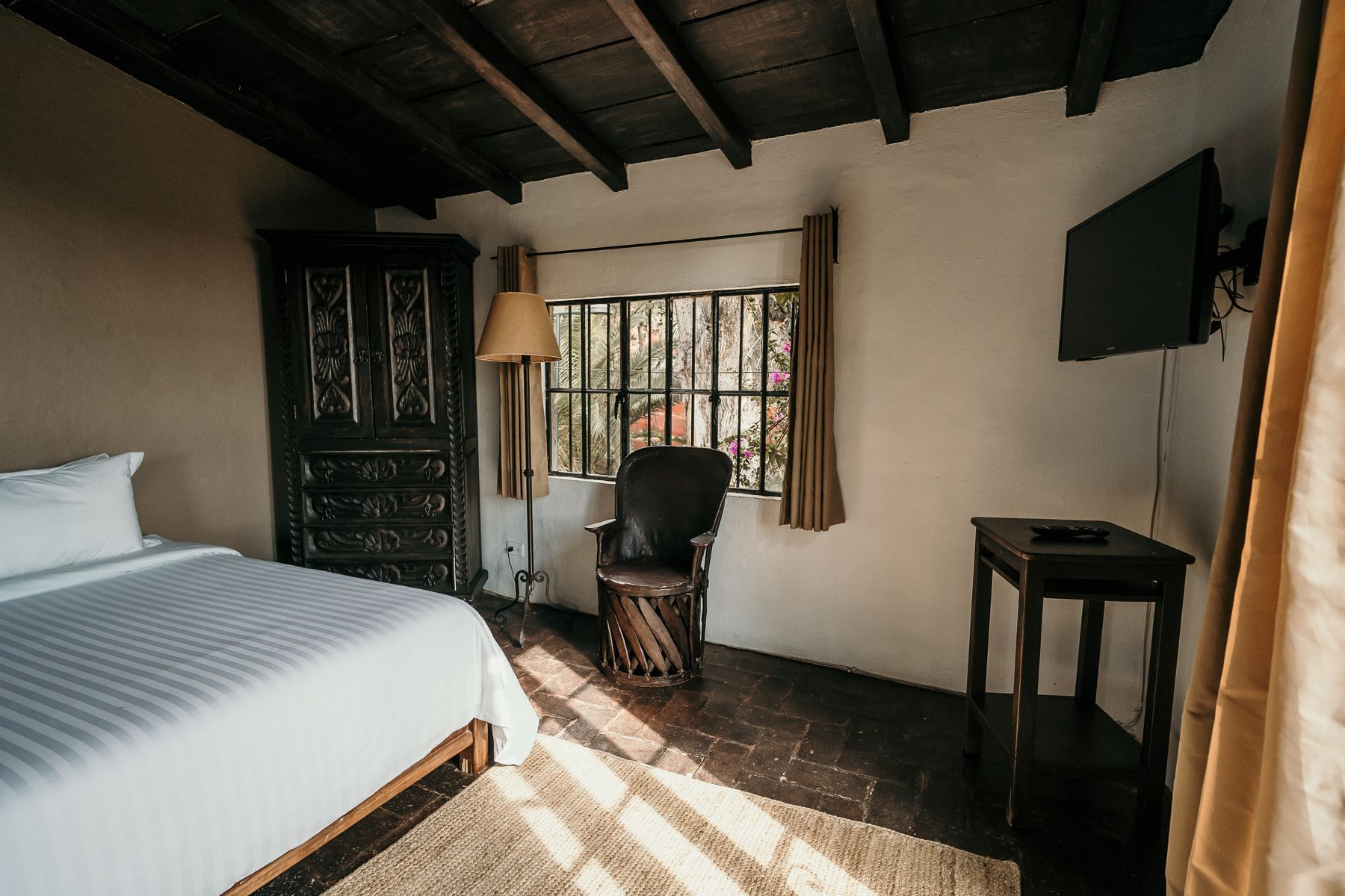 Dormitorio con cama, mueble de madera oscura, silla, ventana, TV y mesa.