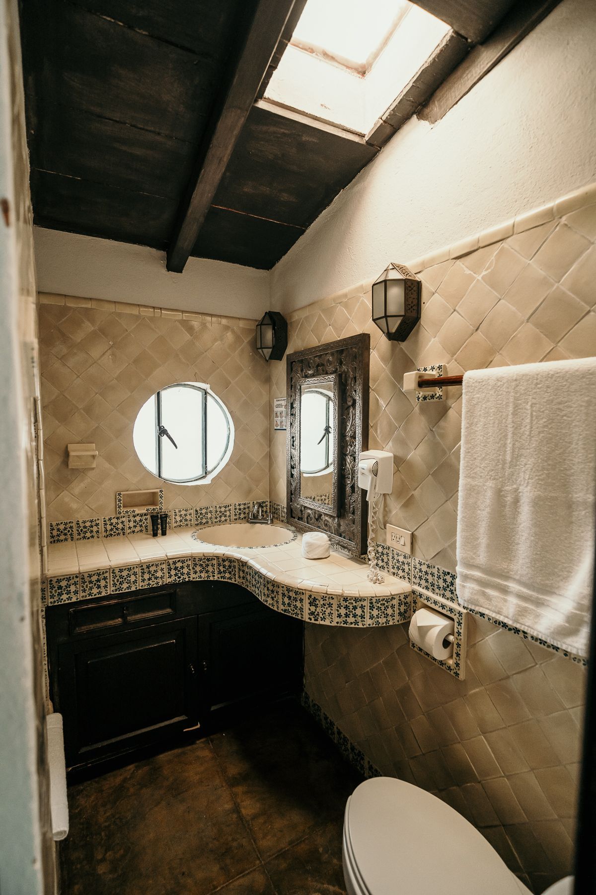 Baño con paredes revestidas de azulejos, tocador de madera oscura, ventana redonda, espejo e inodoro.