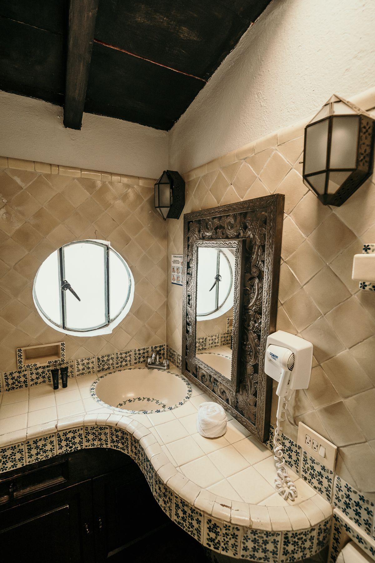 Baño con azulejos estampados, espejo de madera oscura y ventana de ojo de buey.