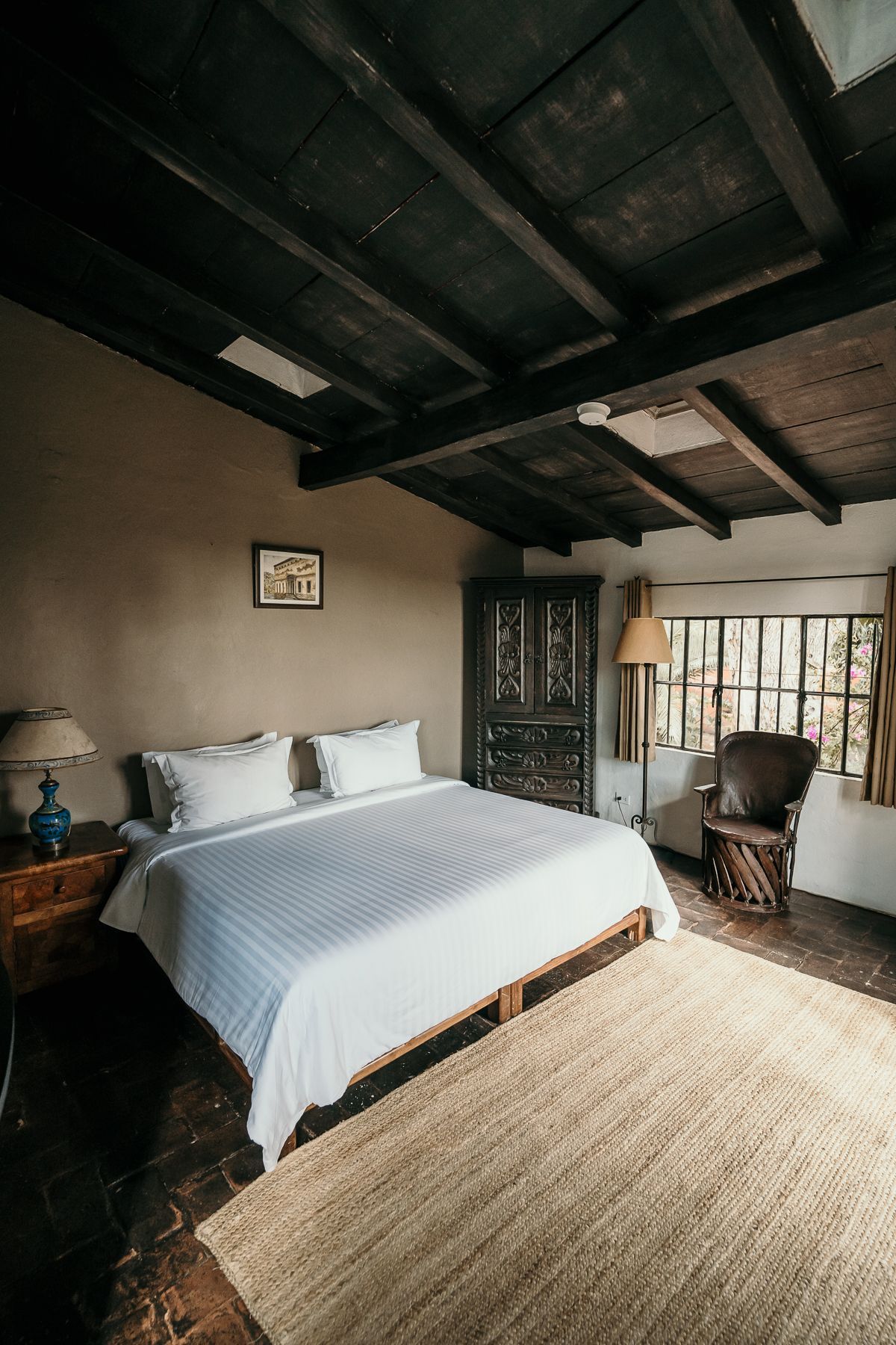 Dormitorio con cama, alfombra, vigas de madera oscura, puerta antigua y ventana con silla.