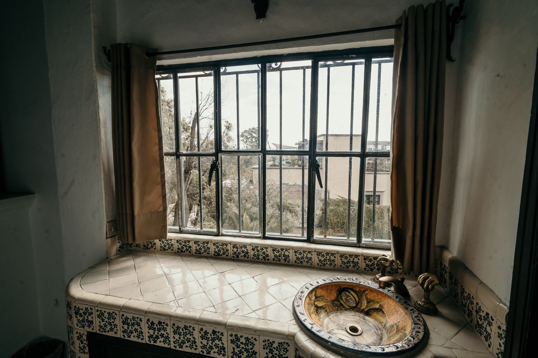 Baño con lavabo debajo de una ventana enrejada, cortinas color canela y encimera estampada.