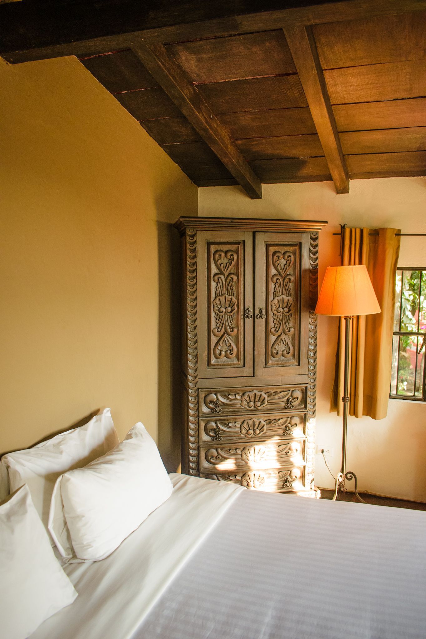 Dormitorio con cama, mueble de madera ornamentado, lámpara de pie y ventana con luz natural.