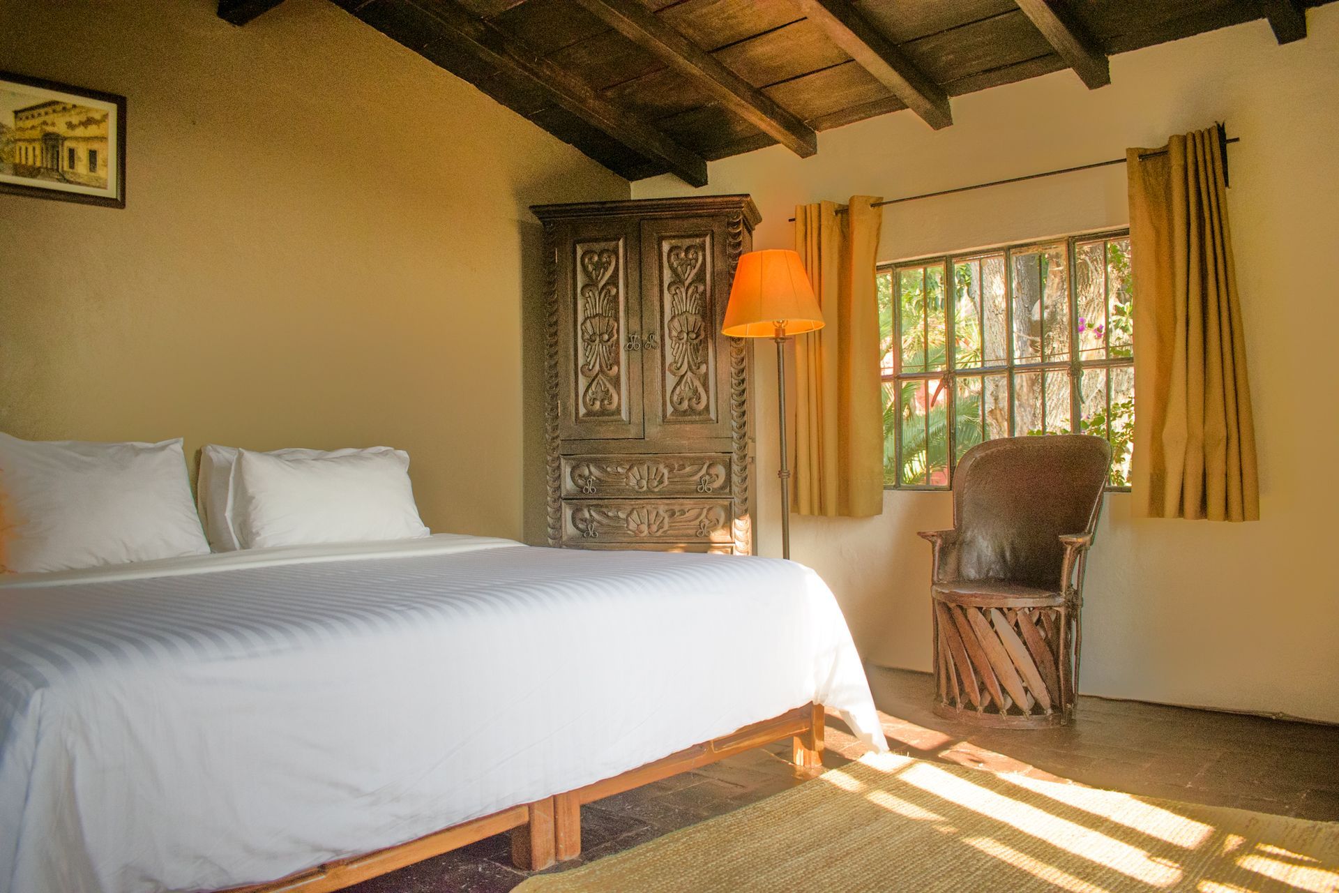 Dormitorio con cama, mueble de madera ornamentado, ventana con luz solar y una silla tejida.