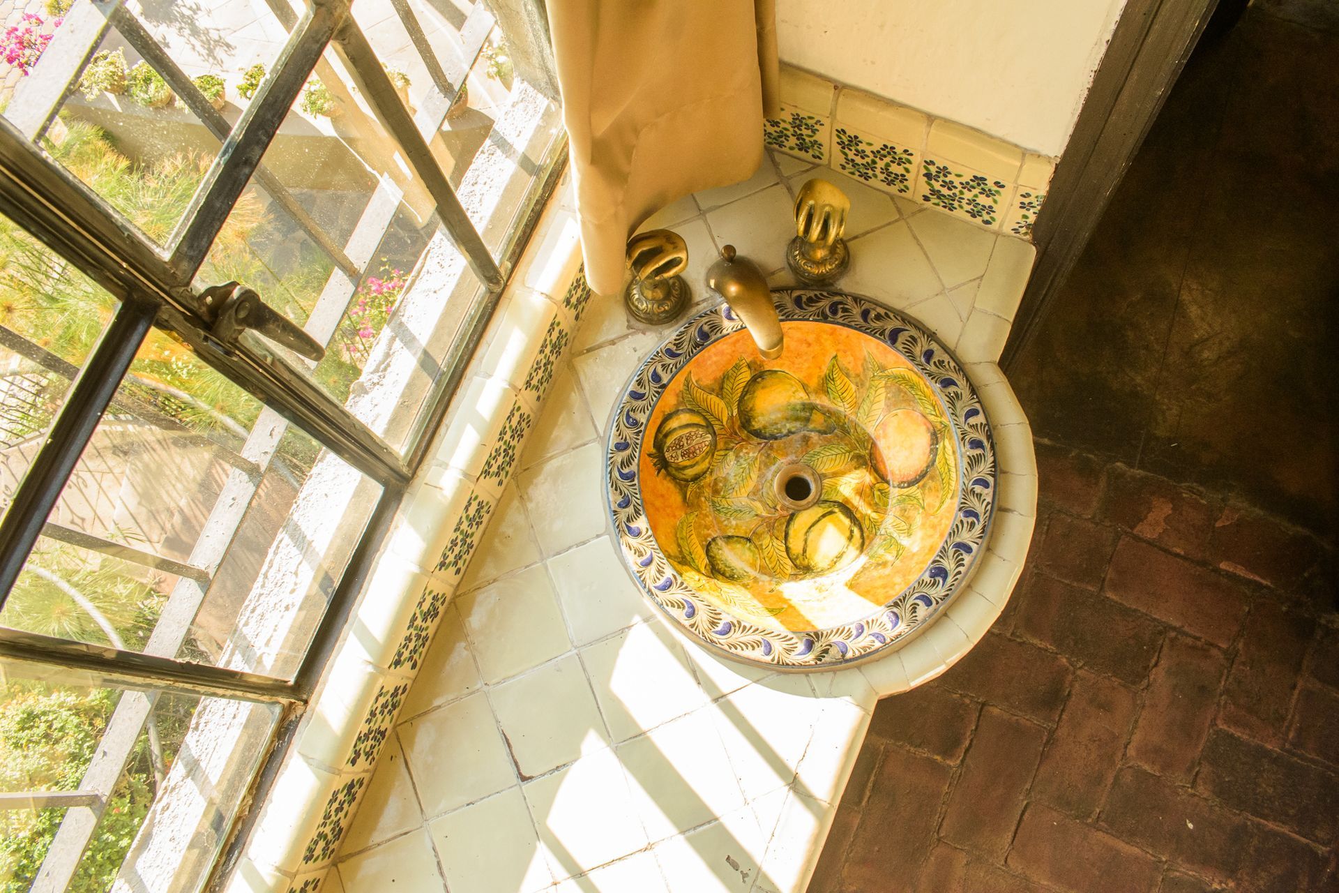 Lavabo decorativo de color amarillo y azul con accesorios dorados junto a una ventana, sobre suelo de baldosas y ladrillo.