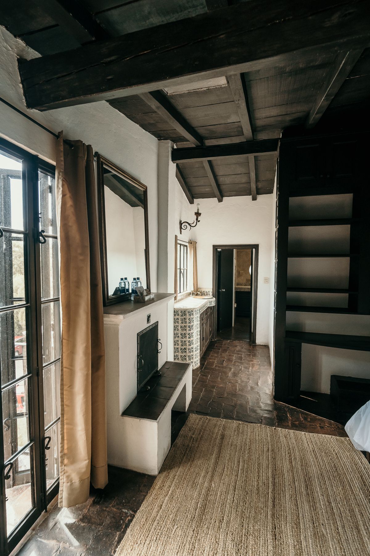 Interior con vigas de madera a la vista, paredes blancas, gran ventanal, alfombra, estanterías empotradas y chimenea.