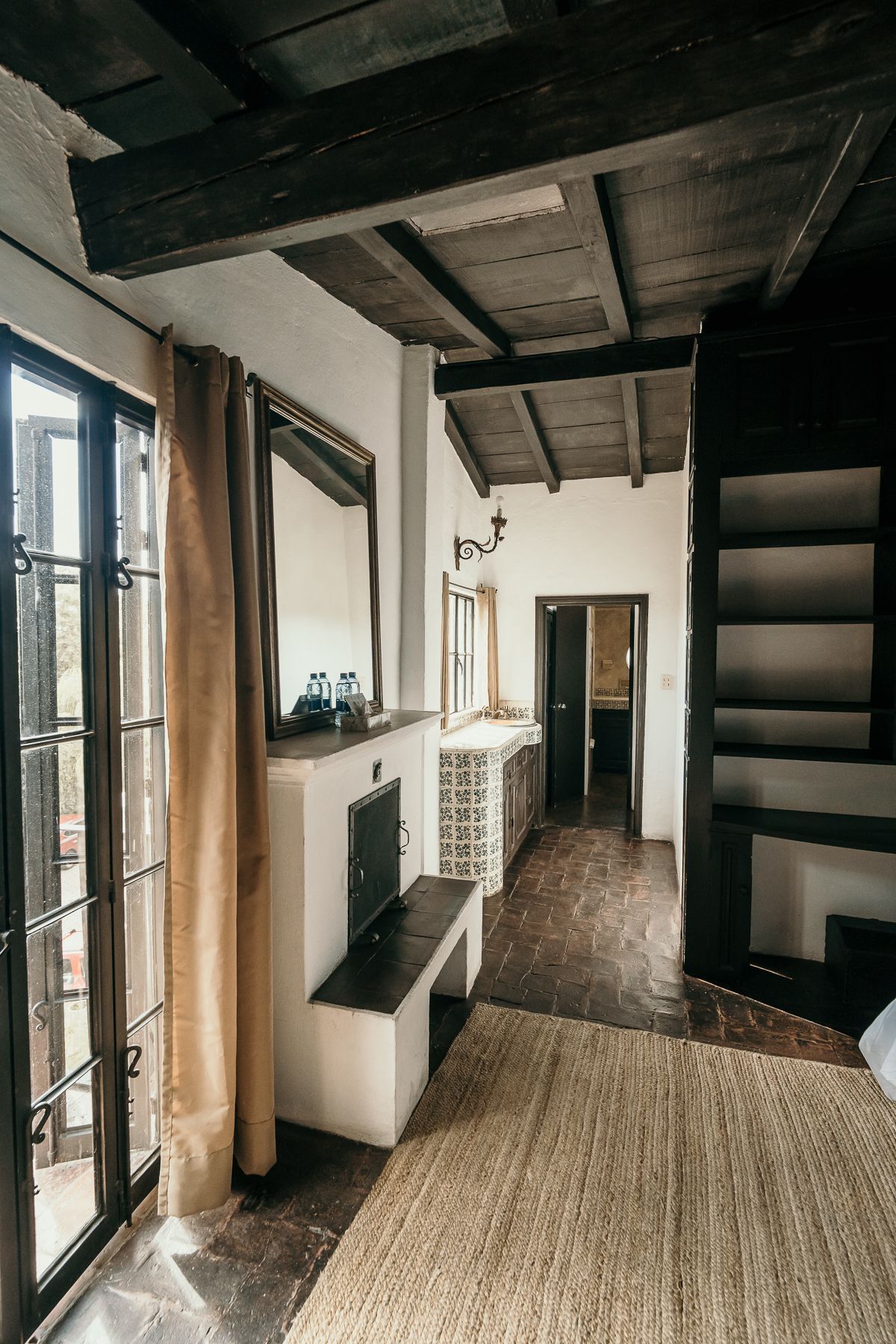 Interior con chimenea, espejo y estanterías empotradas. Una puerta da a un pasillo. Techo con vigas de madera.