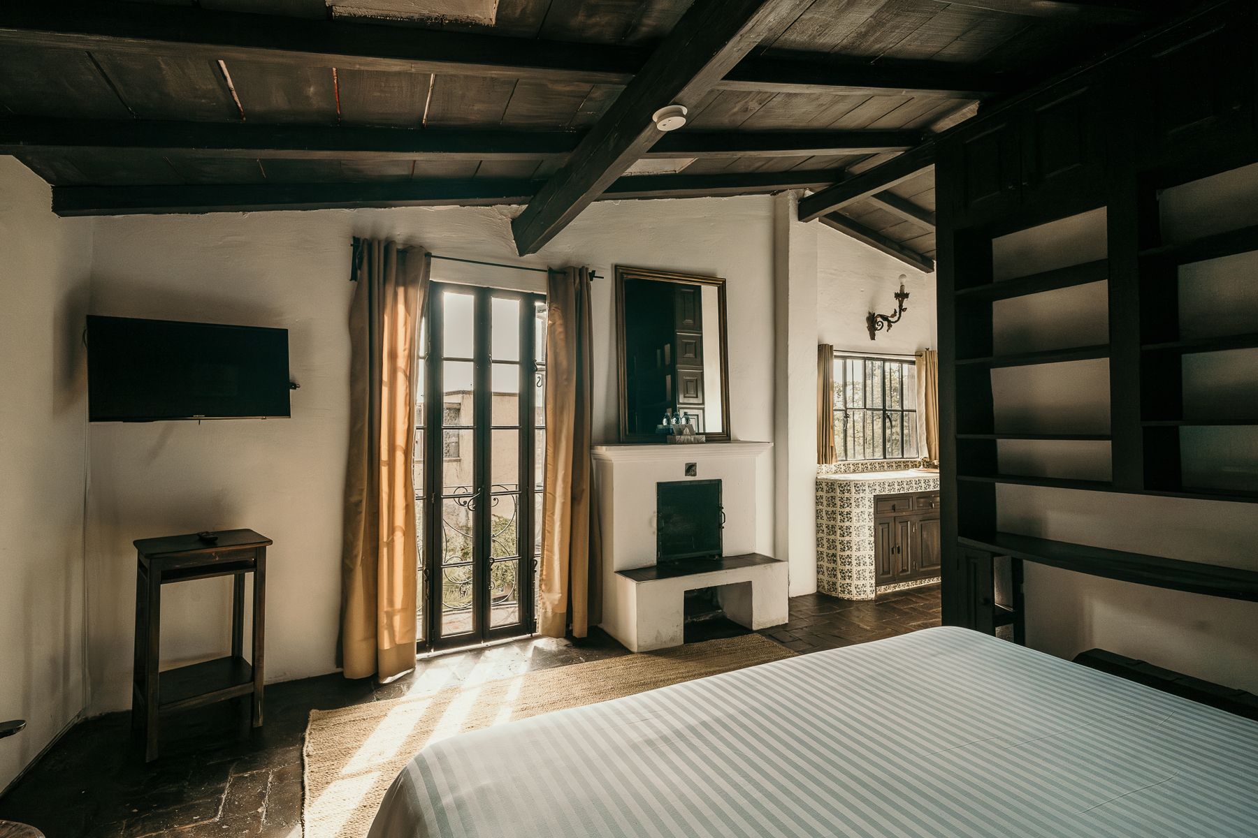 Dormitorio con paredes blancas, techo y vigas de madera oscura, chimenea y puerta con luz solar.