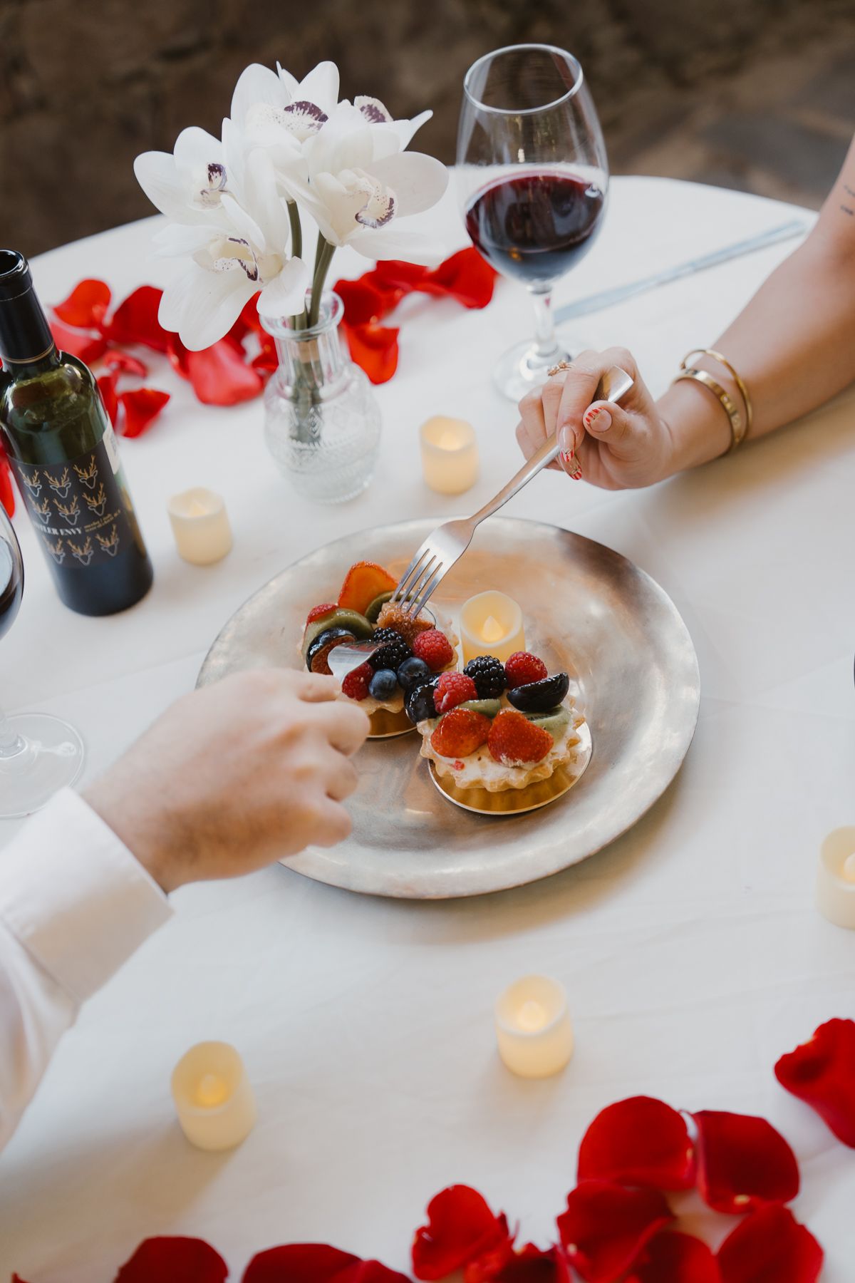 Mesa romántica: pareja compartiendo tartas de frutas, vino y flores, rodeadas de pétalos de rosa y velas.
