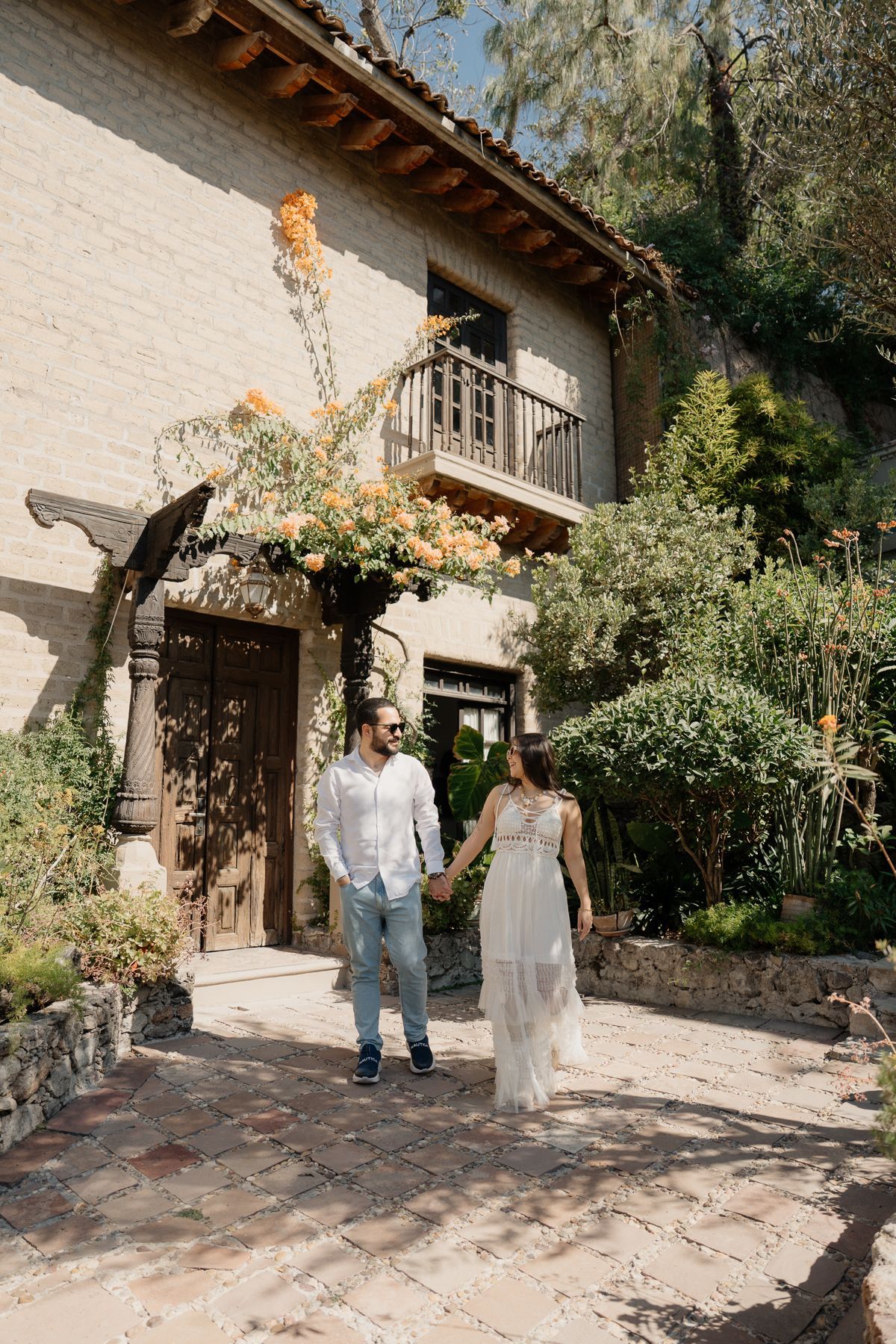 Pareja tomada de la mano, caminando por un camino de ladrillos hacia un edificio de piedra con balcón, rodeado de vegetación.