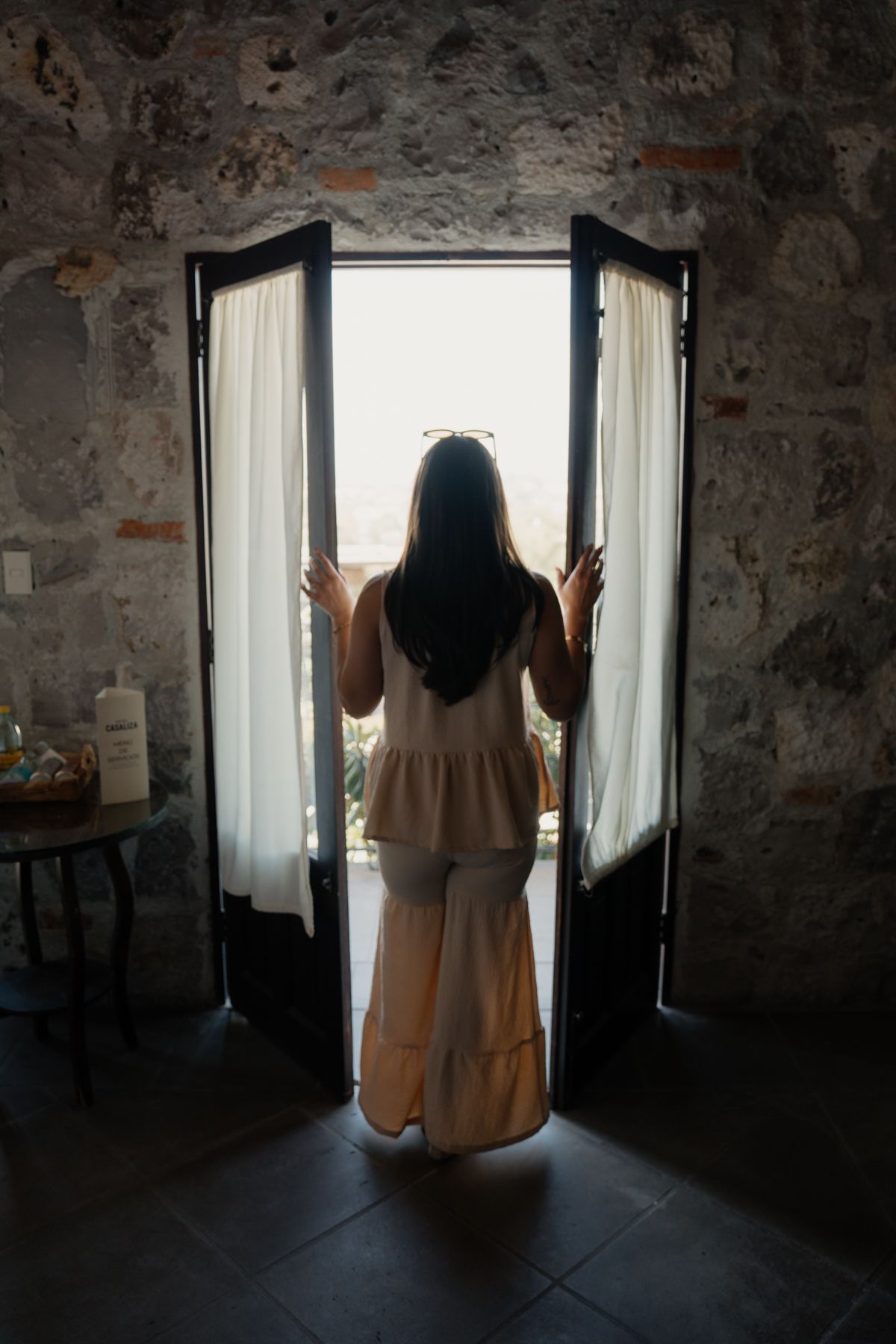 Una mujer está parada en la puerta, mirando hacia afuera. La luz entra a raudales. Paredes de piedra.
