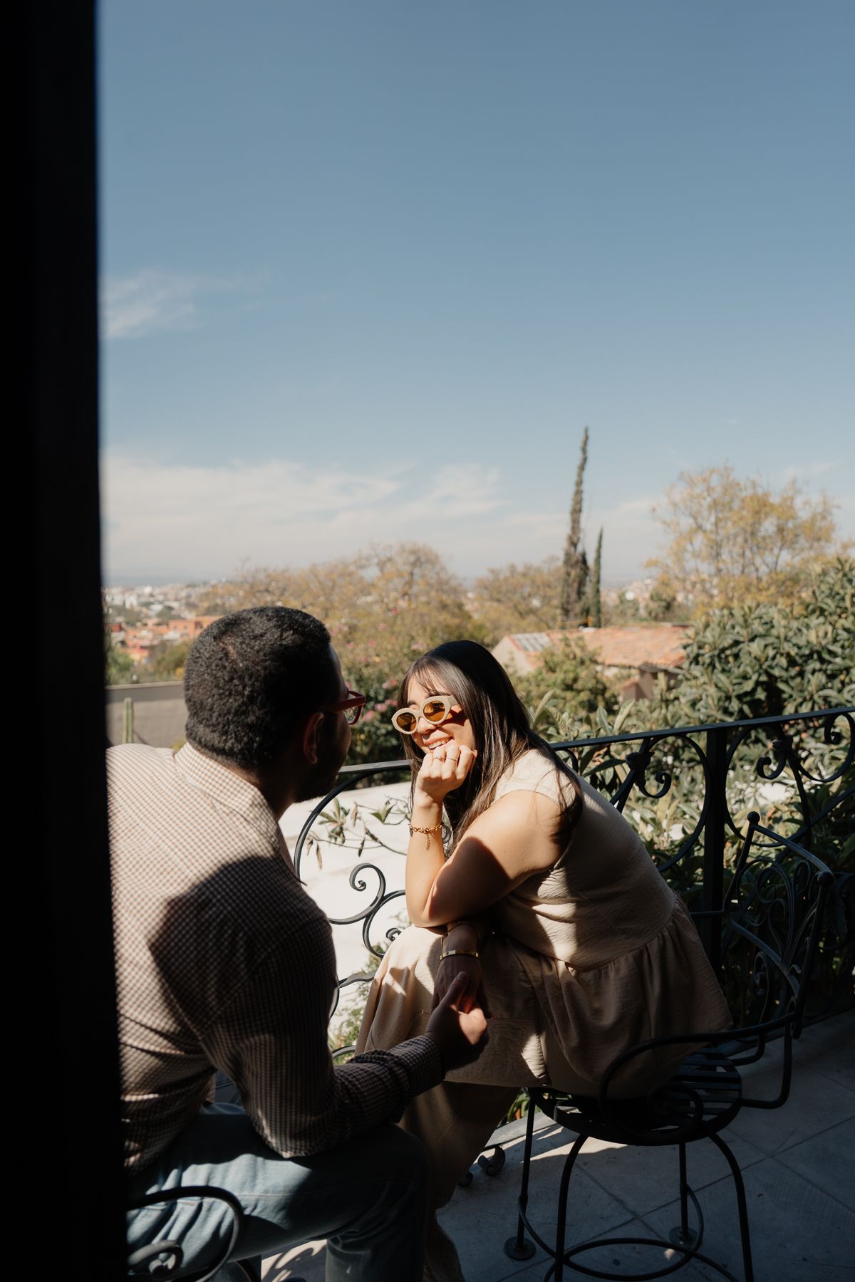 Pareja sentada en un balcón, un día soleado. La mujer sonríe, mirando al hombre. Árboles y paisaje urbano al fondo.