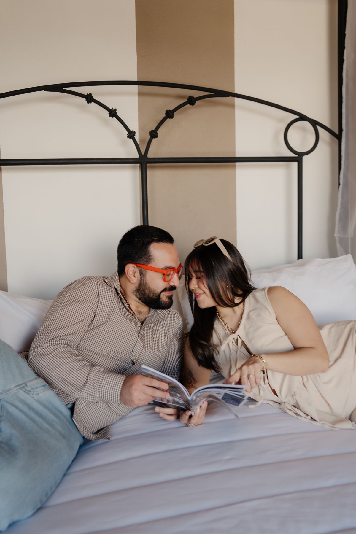 Pareja en una cama mirando un libro; el hombre lleva gafas y camisa estampada; la mujer lleva un vestido y una diadema.