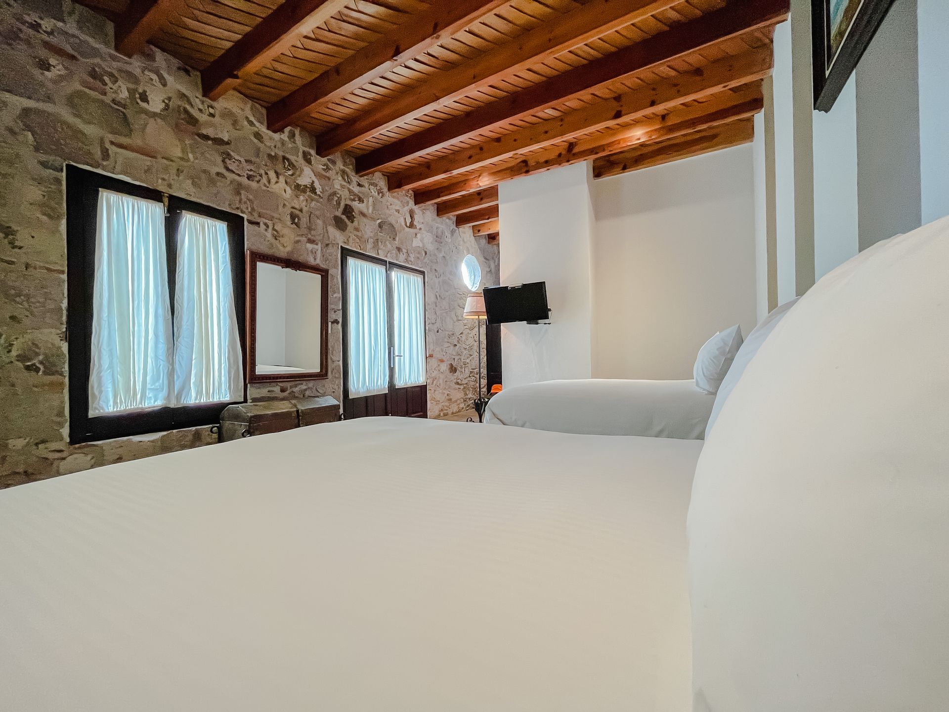 Dormitorio con pared de piedra, techo de madera, dos ventanas con cortinas blancas, cama blanca.